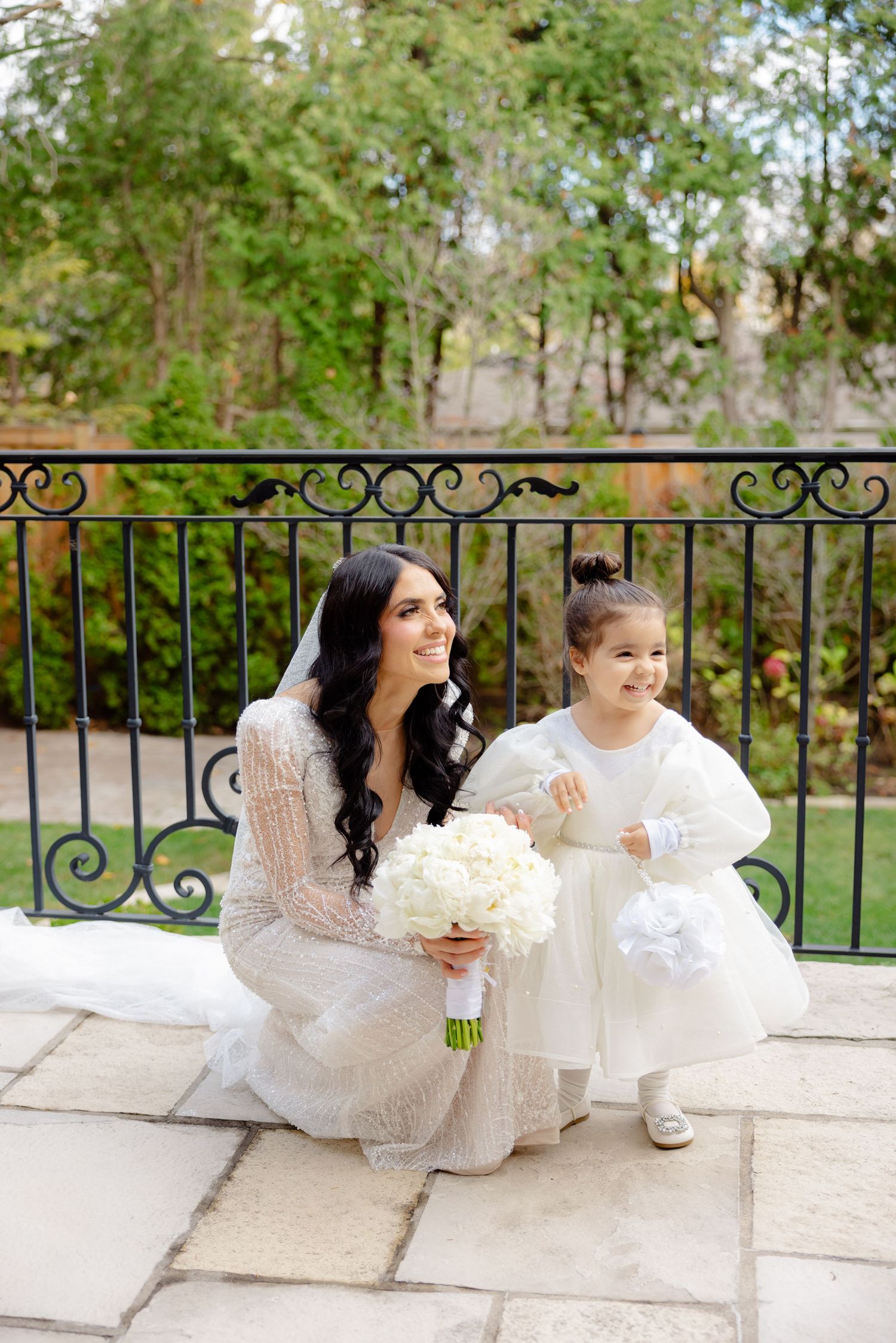 Elegant bride in lace gown kneels beside flower girl on stone terrace with garden backdrop.