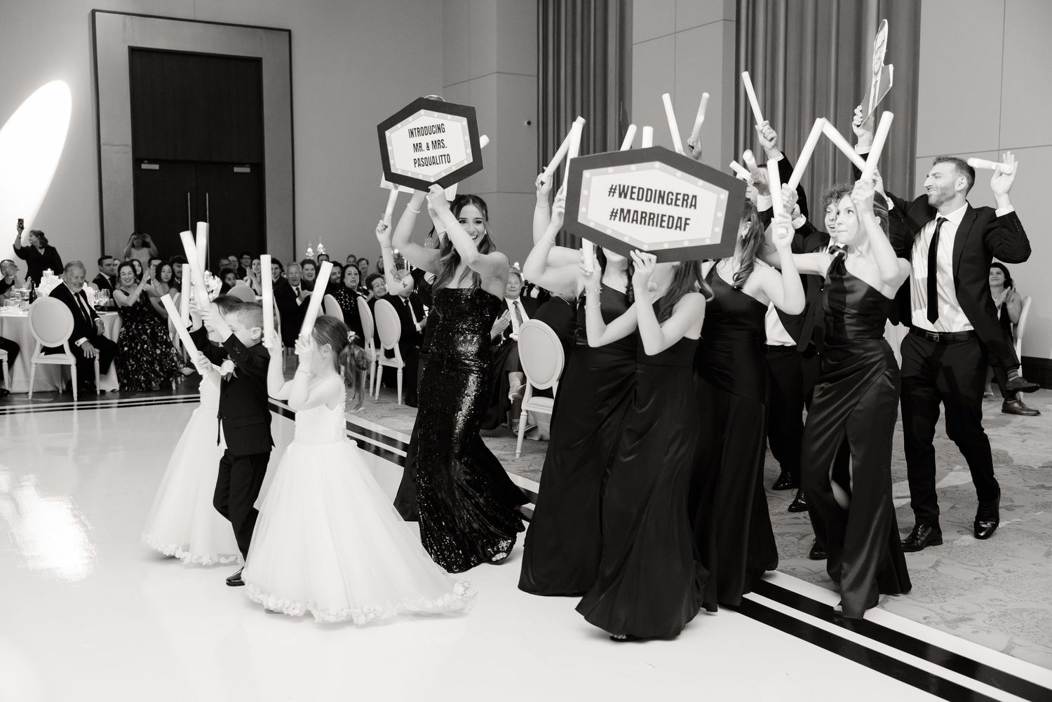 Wedding reception guests holding up fun celebration signs while dancing in black and white formal attire on dance floor.
