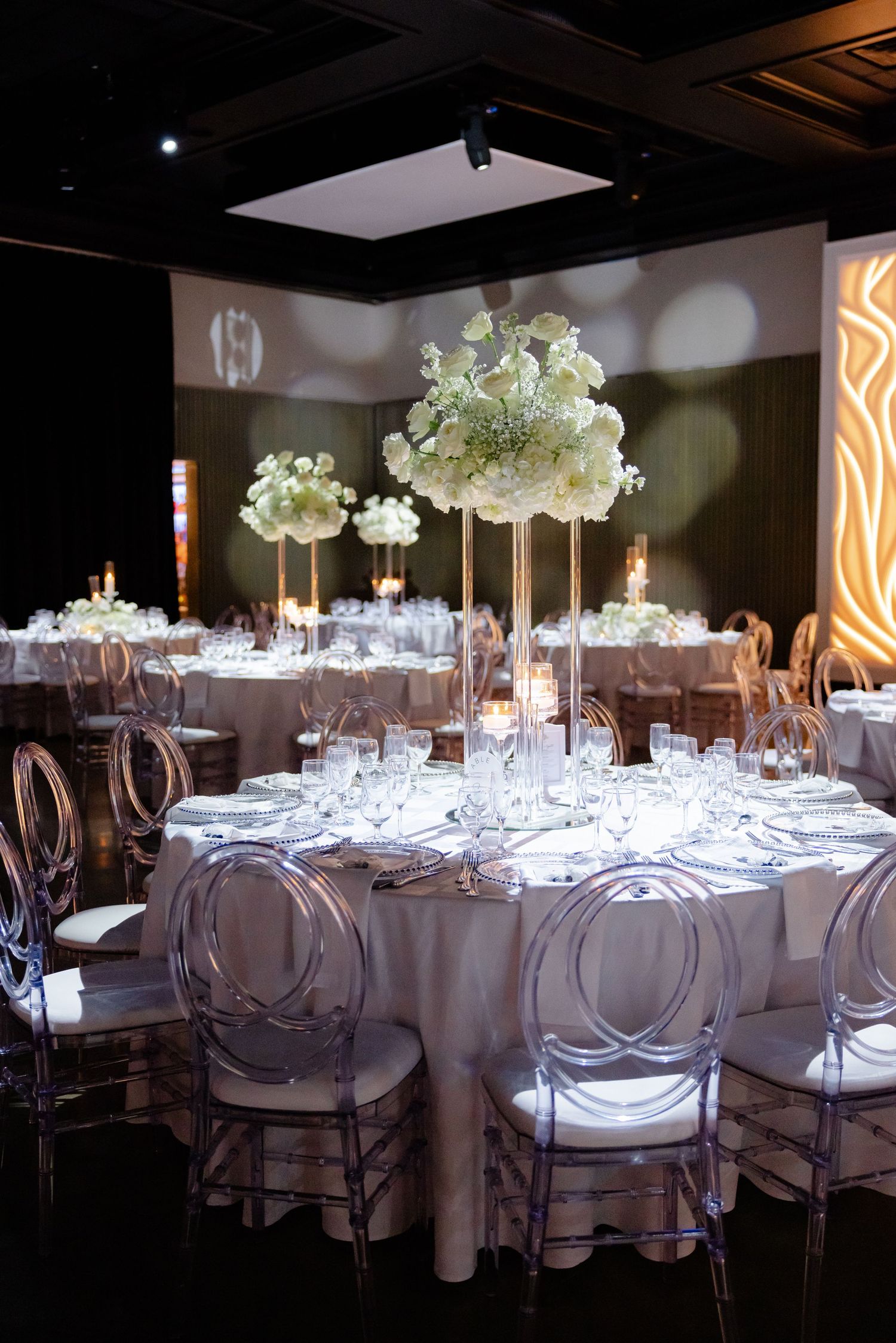 Elegant wedding reception tables with tall white floral centerpieces and ghost chairs in modern venue space.