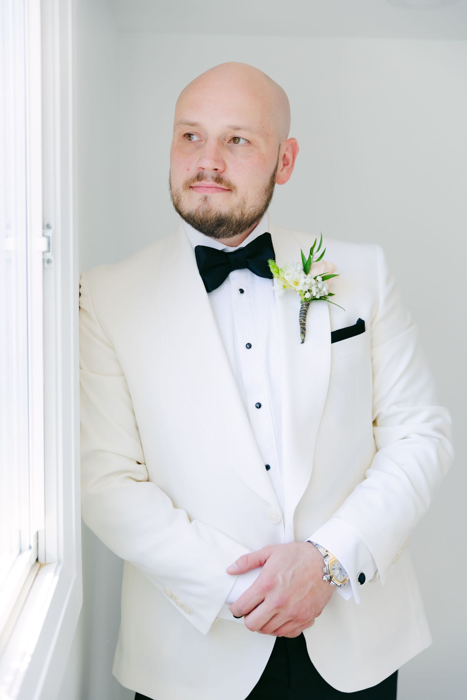 Elegant groom in white tuxedo jacket and black bow tie with boutonniere standing by window.