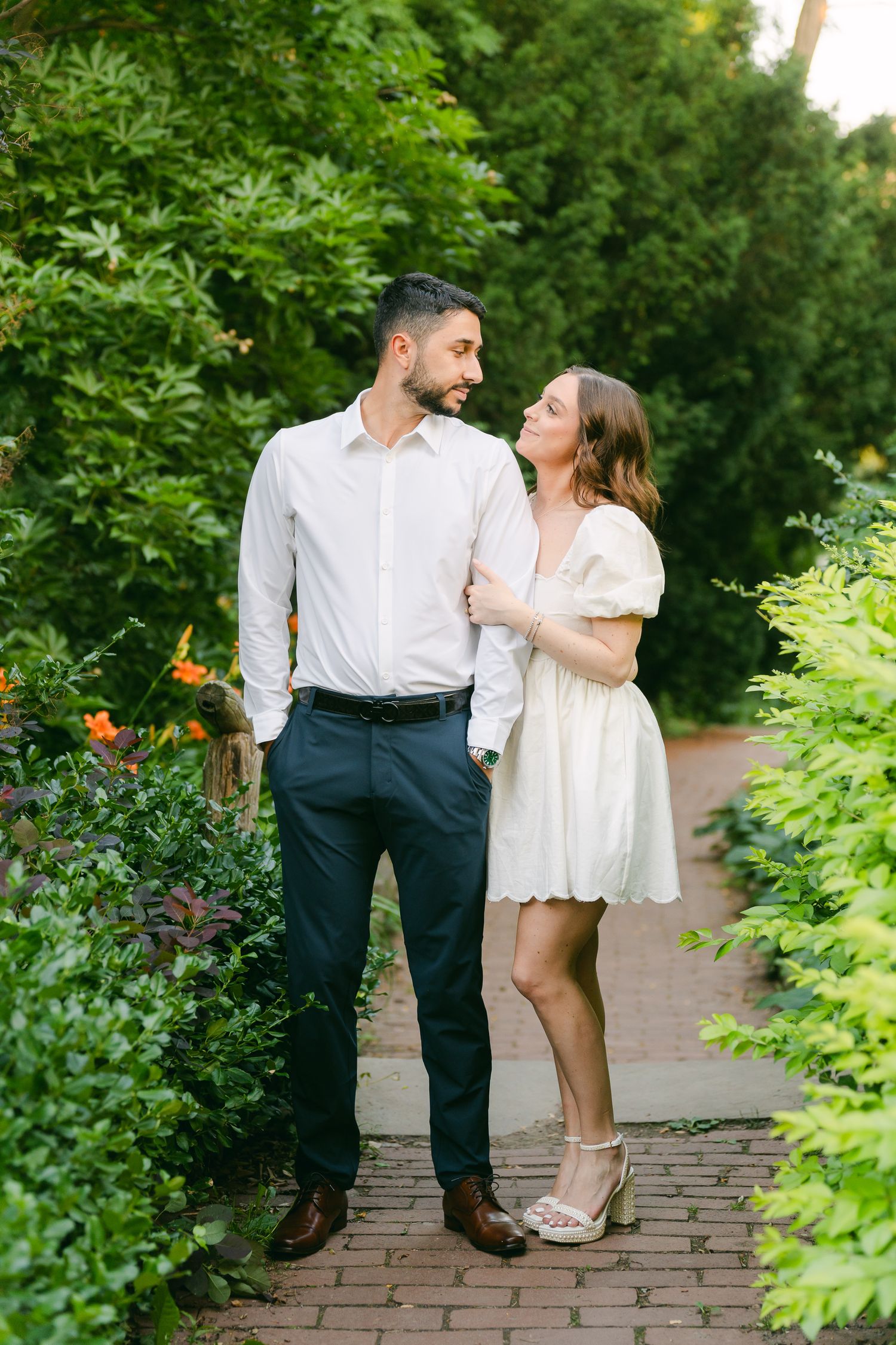 Couple exchanging loving glances while standing on brick path amid lush garden scenery.