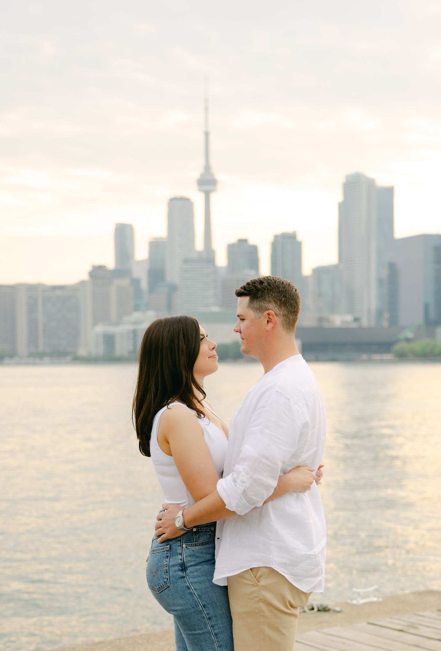 Romantic couple embraces by Toronto waterfront at sunset with CN Tower skyline in background.