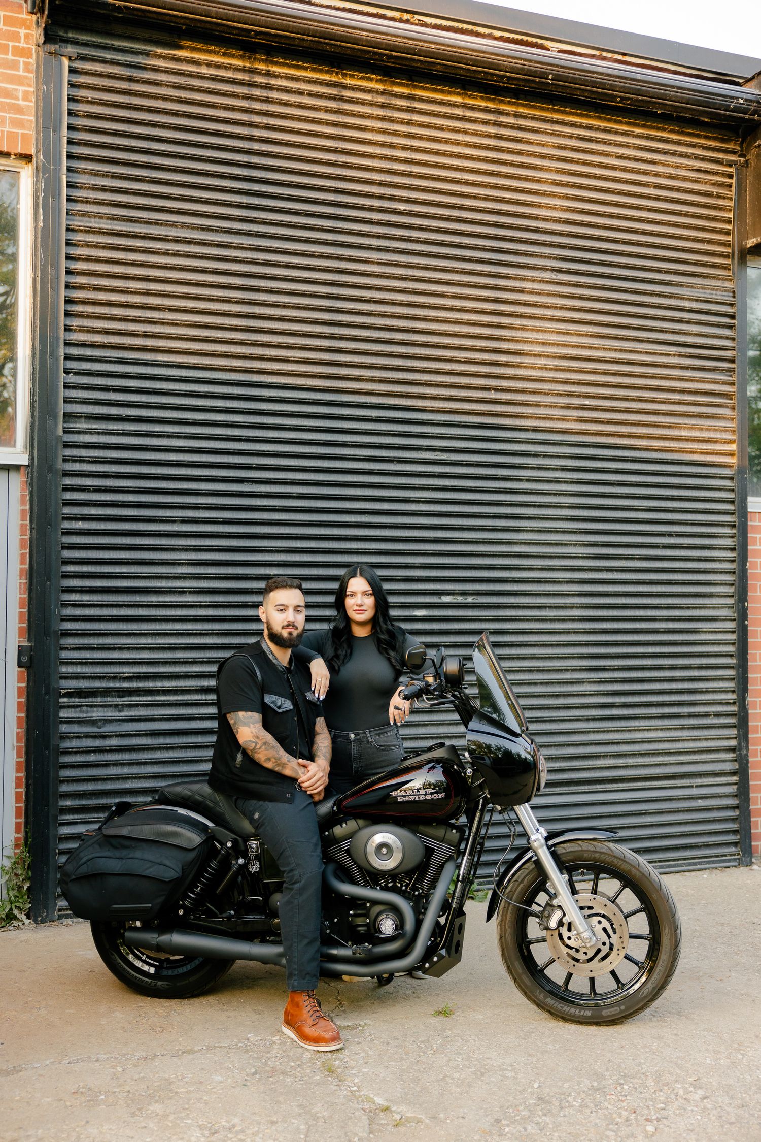A couple poses with their custom black motorcycle against a metal garage door background.