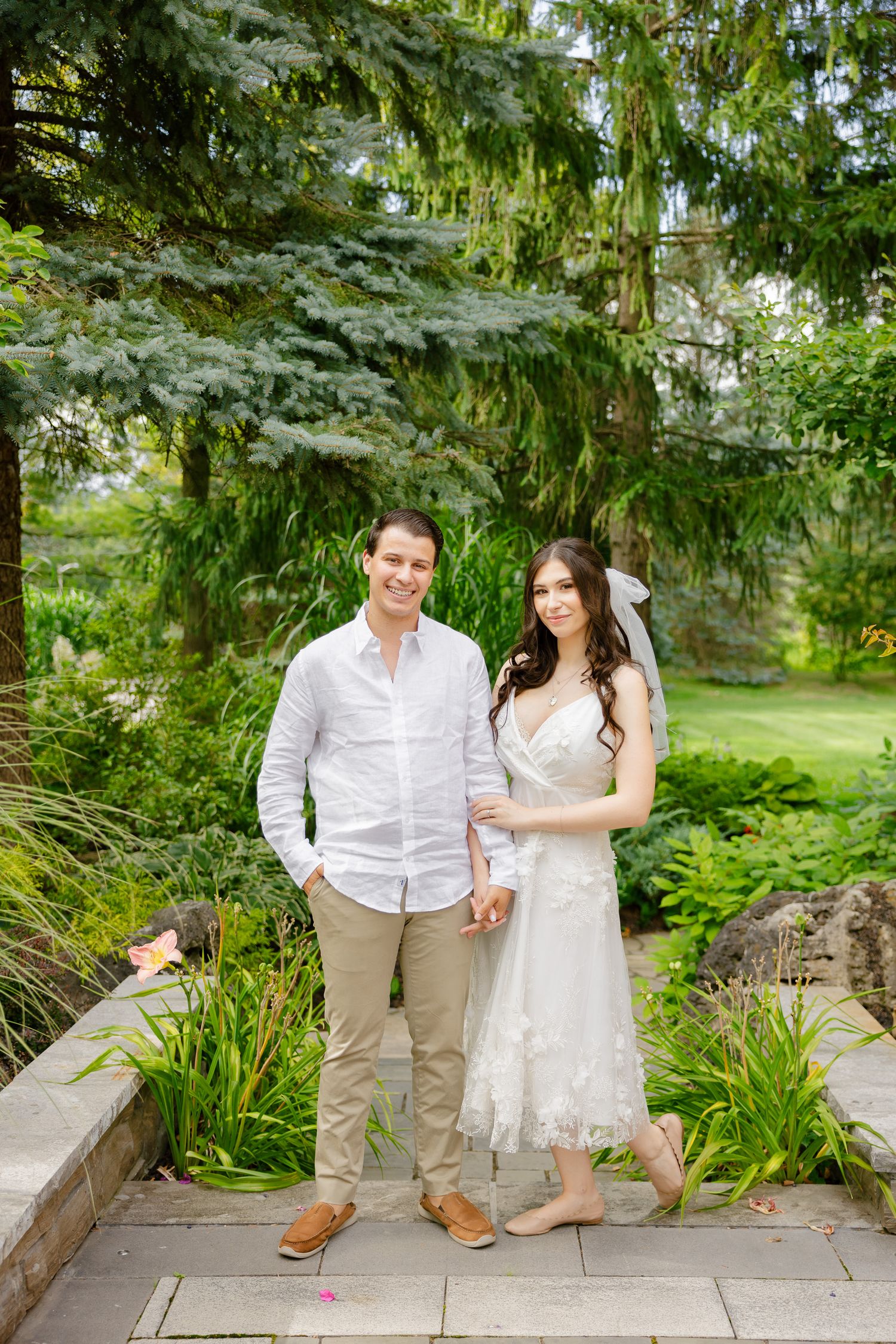 Casual wedding portrait on stone pathway surrounded by lush garden greenery.