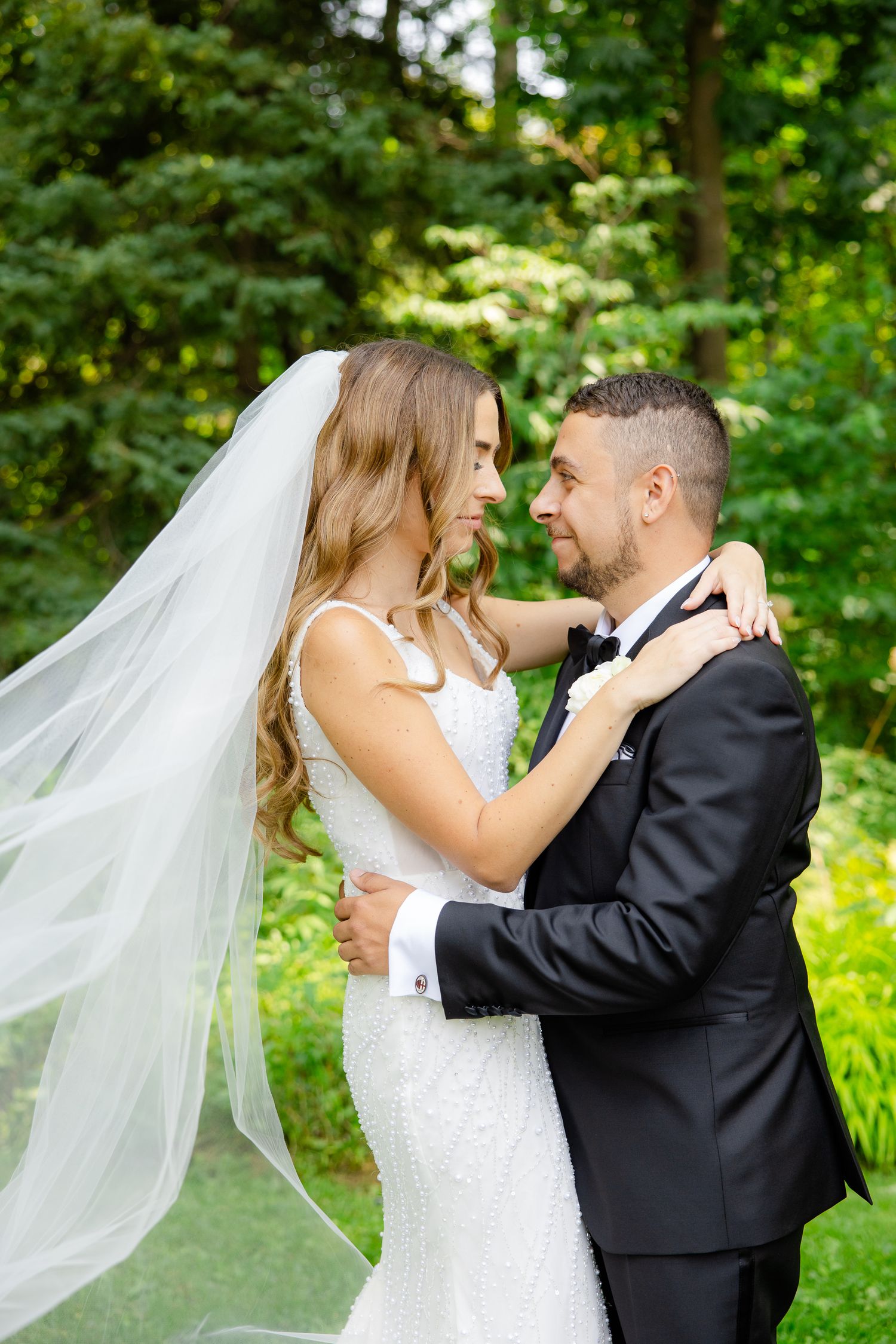 Romantic outdoor portrait of wedding couple embracing with flowing veil and forest backdrop.