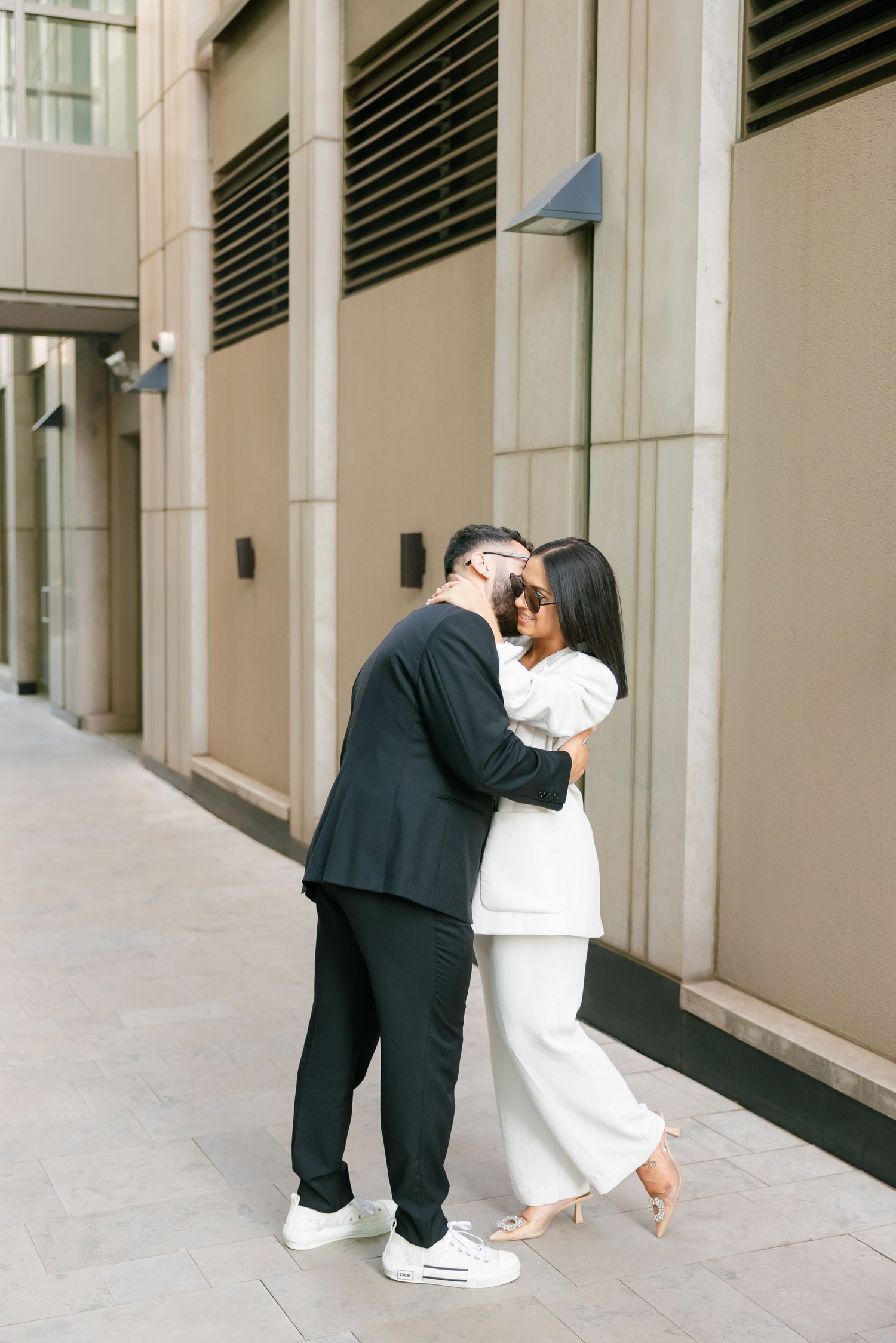 A romantic couple in formal attire share an intimate moment against a beige wall.