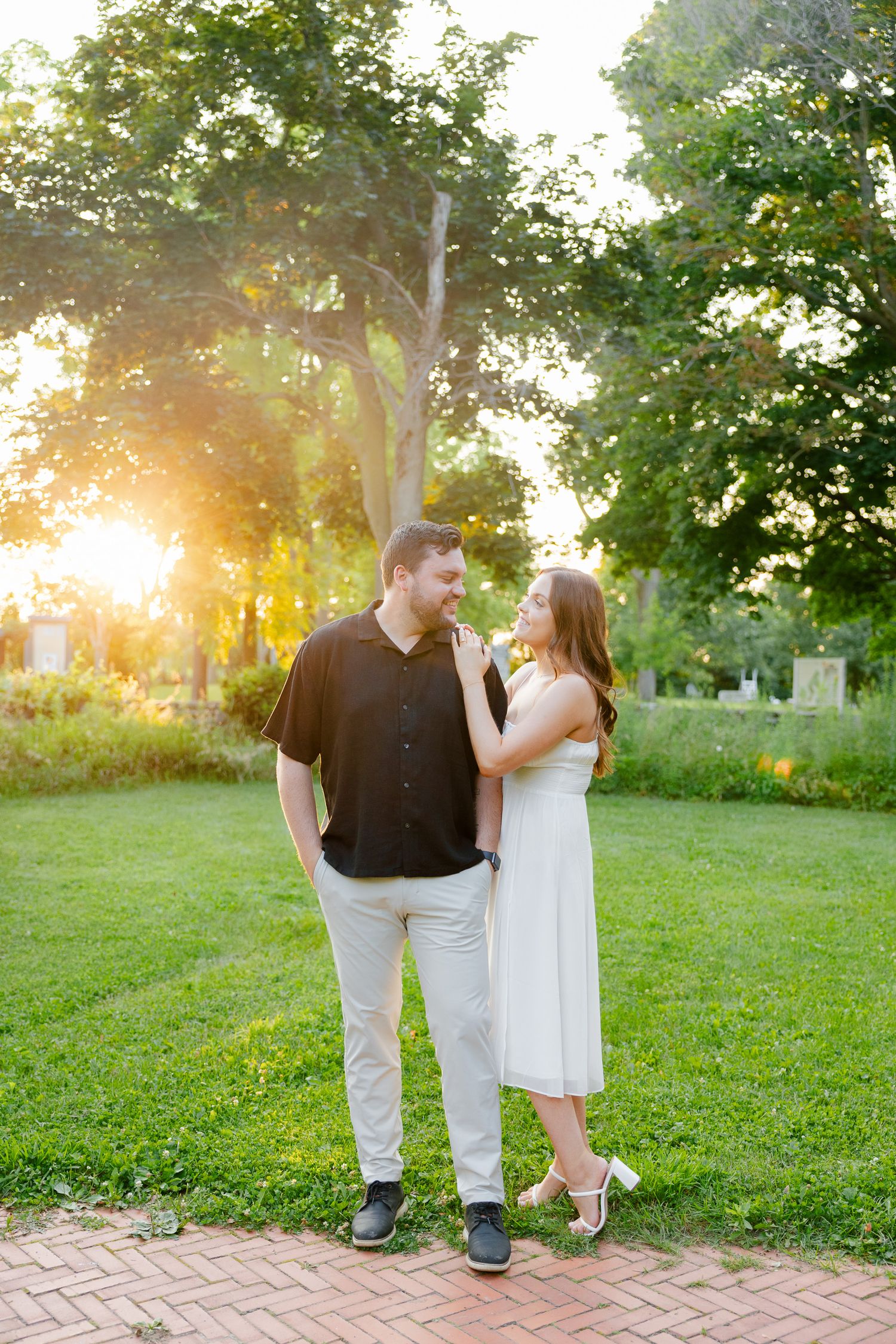 Couple in contrasting outfits stand together on brick path with glowing sunset behind trees.