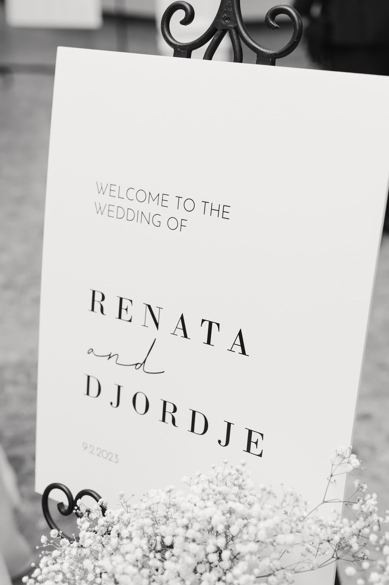Black and white wedding welcome sign featuring minimalist typography and ornate metal stand detail.