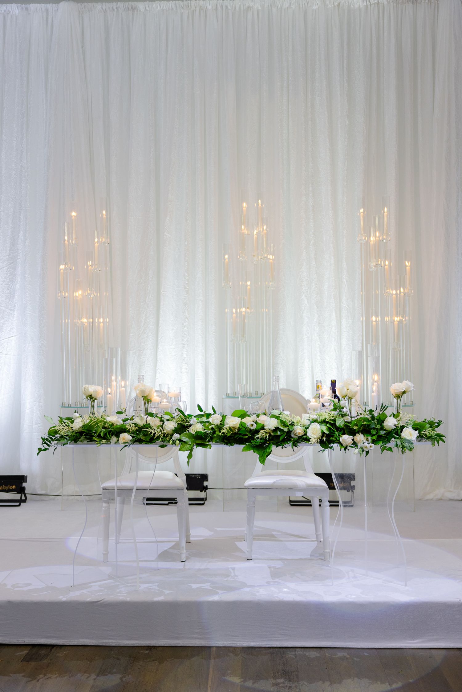 Minimalist white wedding backdrop with tiered floral arrangements and glowing candle displays.