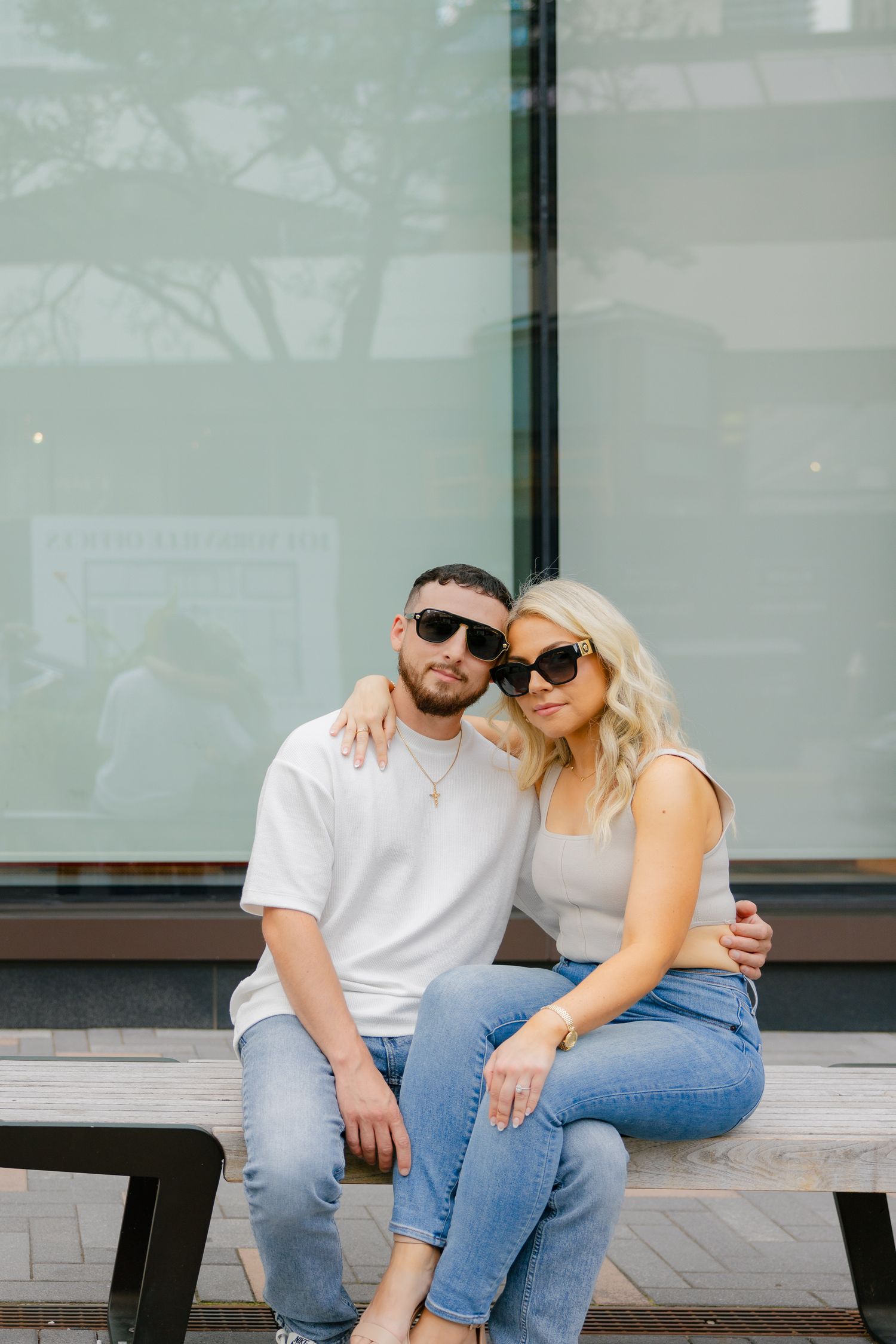 Couple in matching sunglasses embrace while sitting on bench outside storefront window.