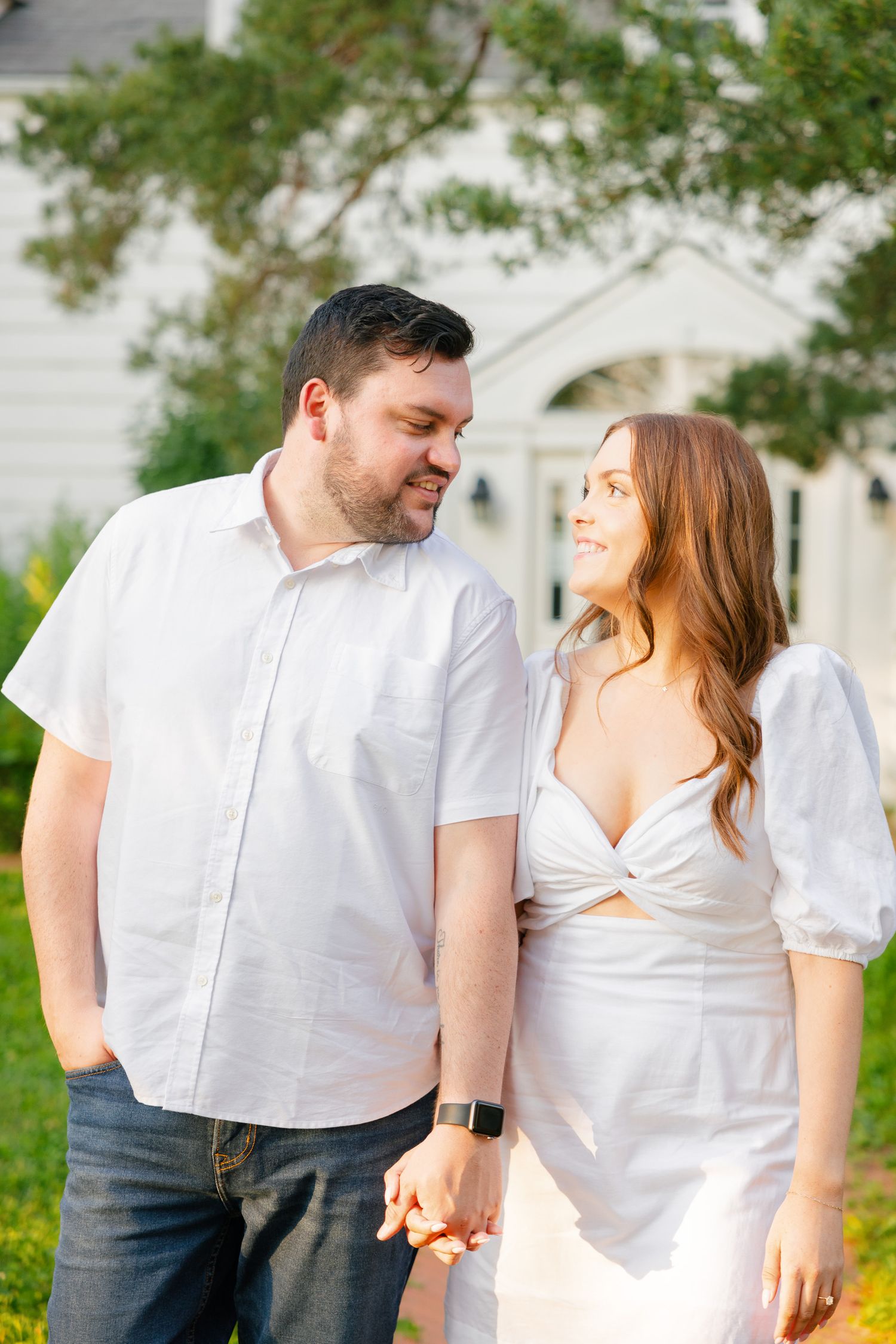 A couple in white outfits shares a loving glance while holding hands in a garden setting.