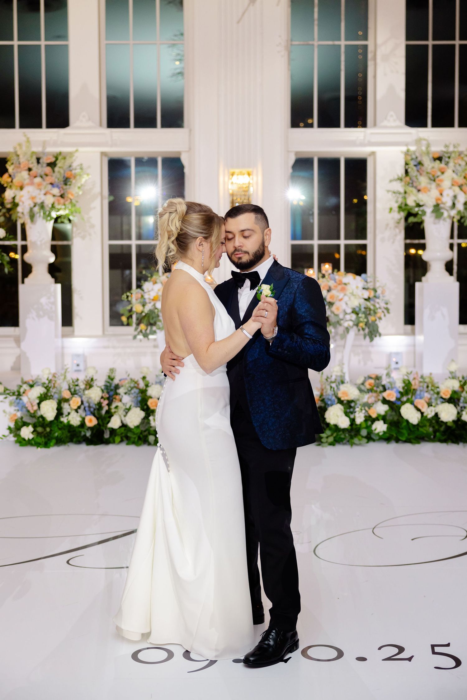 Couple dancing together in an elegant white ballroom with floral decorations and large windows.