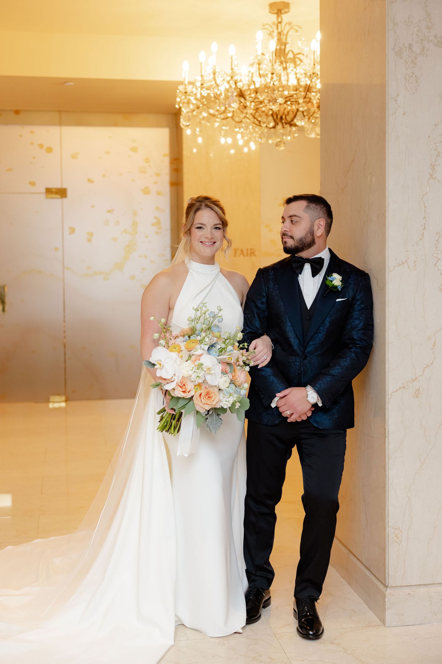 A bride and groom share a candid moment in a marble-walled venue with chandeliers overhead.