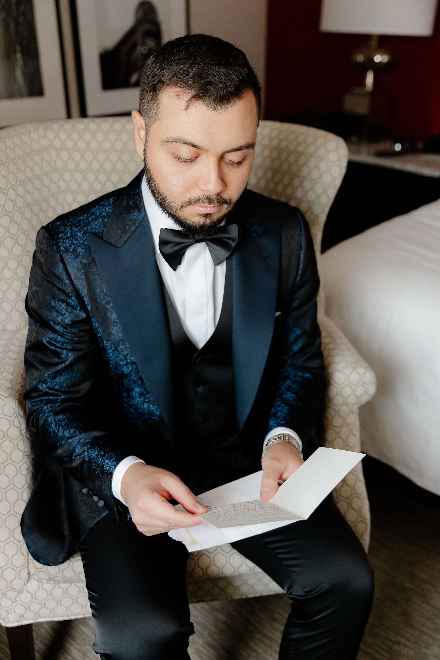 A person in a textured navy tuxedo reads a document while seated in an upholstered chair.