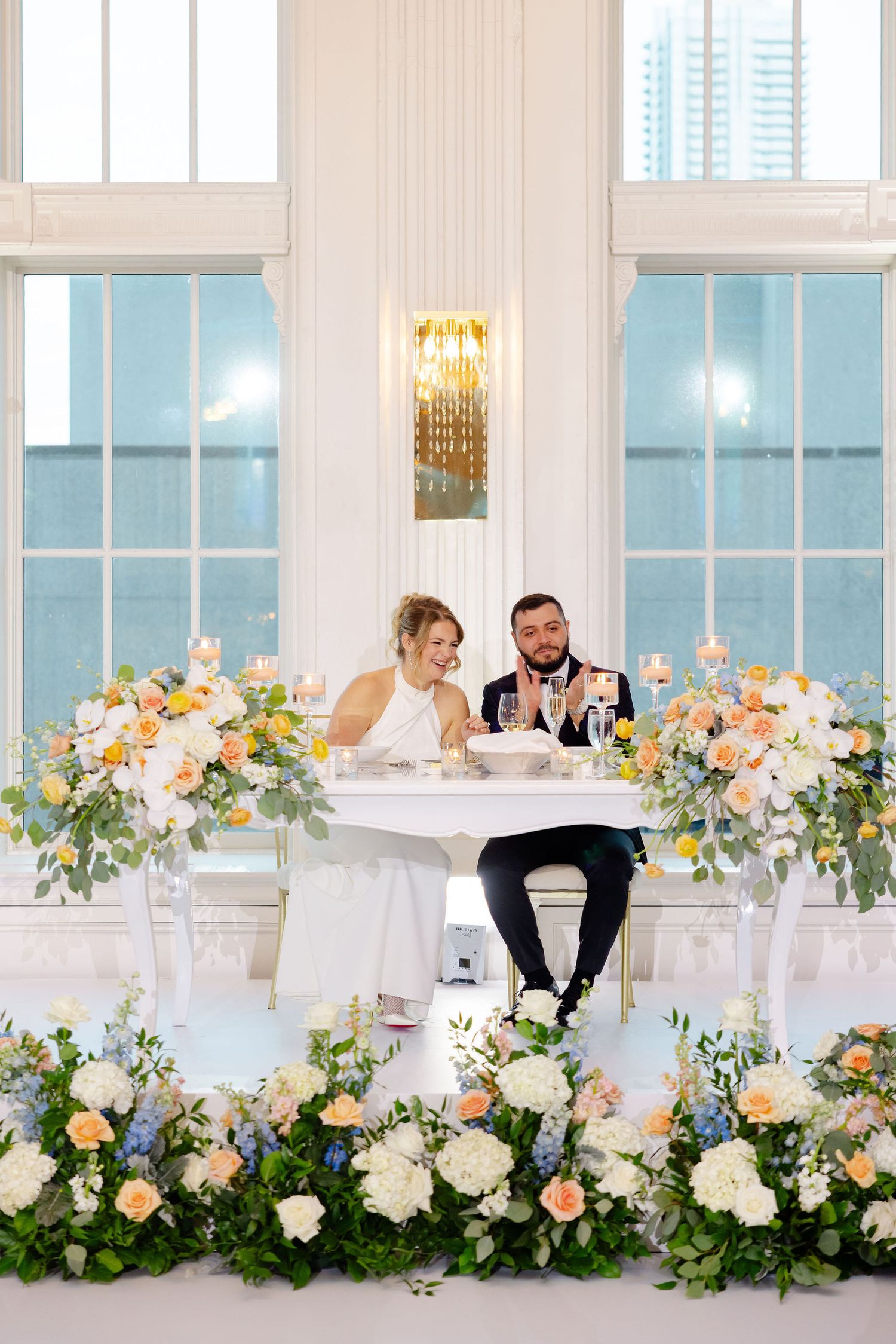Elegant wedding reception table decorated with lush floral arrangements in peach and white with greenery at a bright venue.