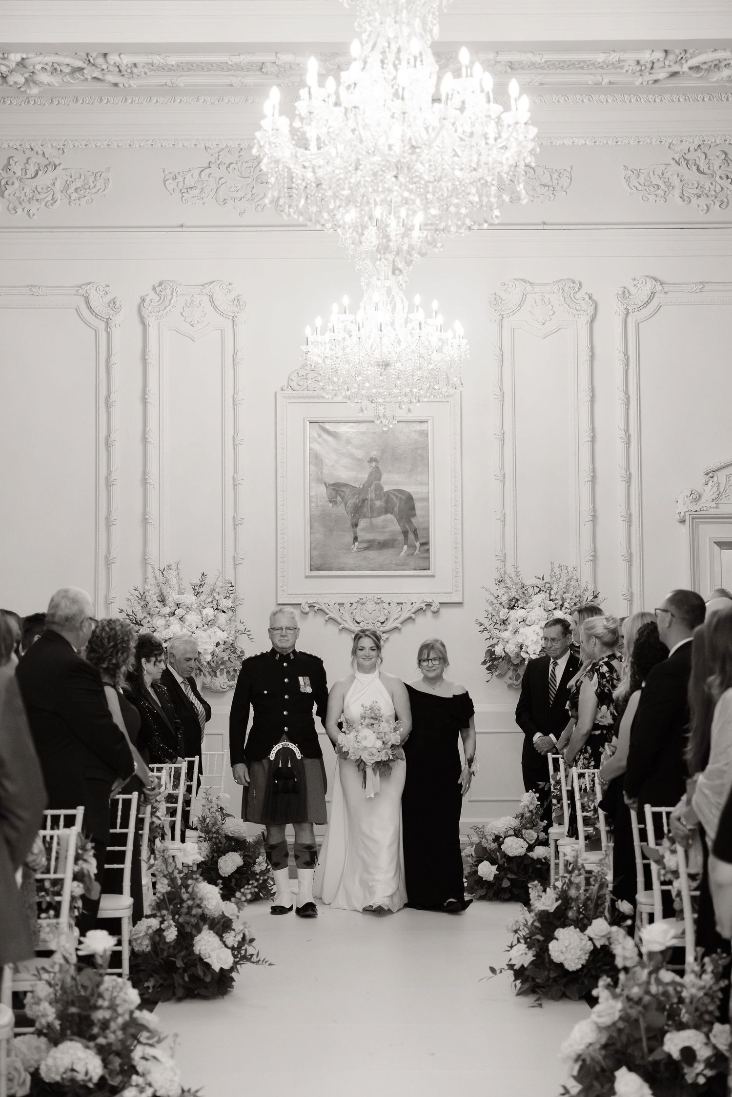 Wedding ceremony setup in grand white room with crystal chandeliers and elegant floral arrangements.