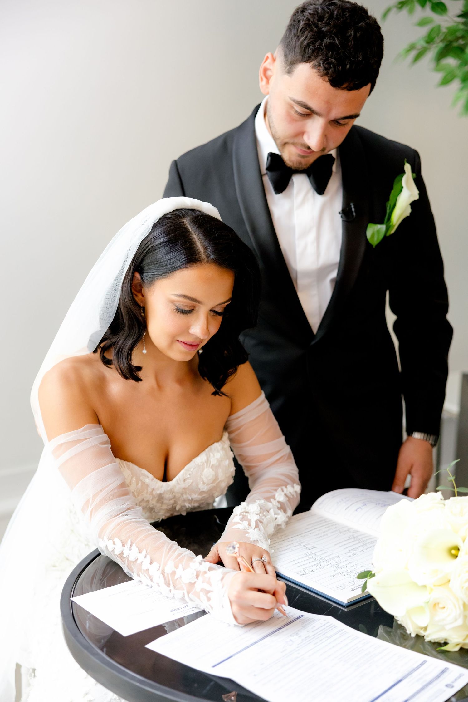 Couple signing marriage documents at small table with white floral arrangements.