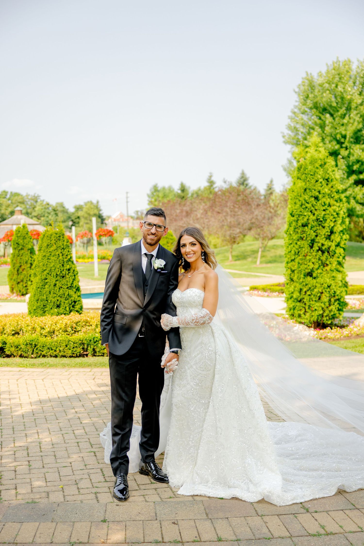 Wedding couple poses together on brick path surrounded by manicured evergreen trees and landscaping.