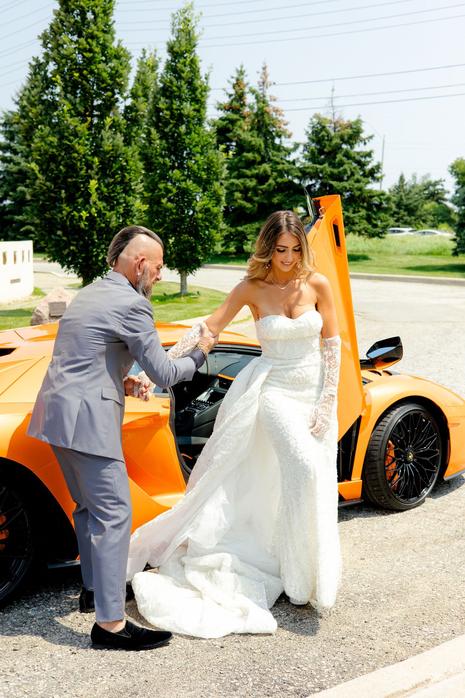 A bride in a white gown exits an orange Lamborghini while being assisted on a sunny day.