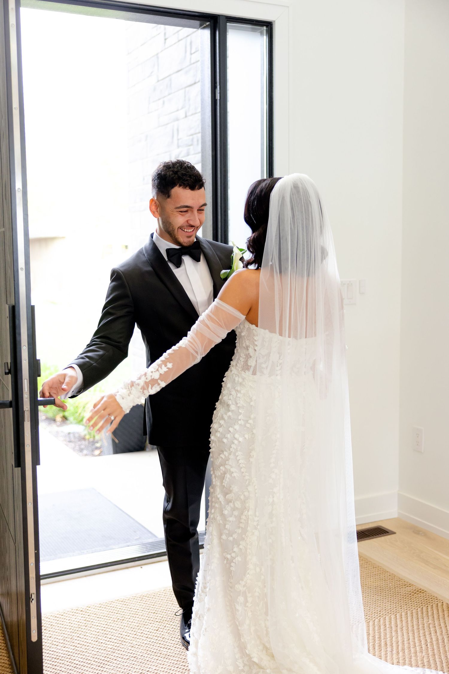 A bride in a lace gown and veil stands with her groom wearing a black tuxedo by a doorway.