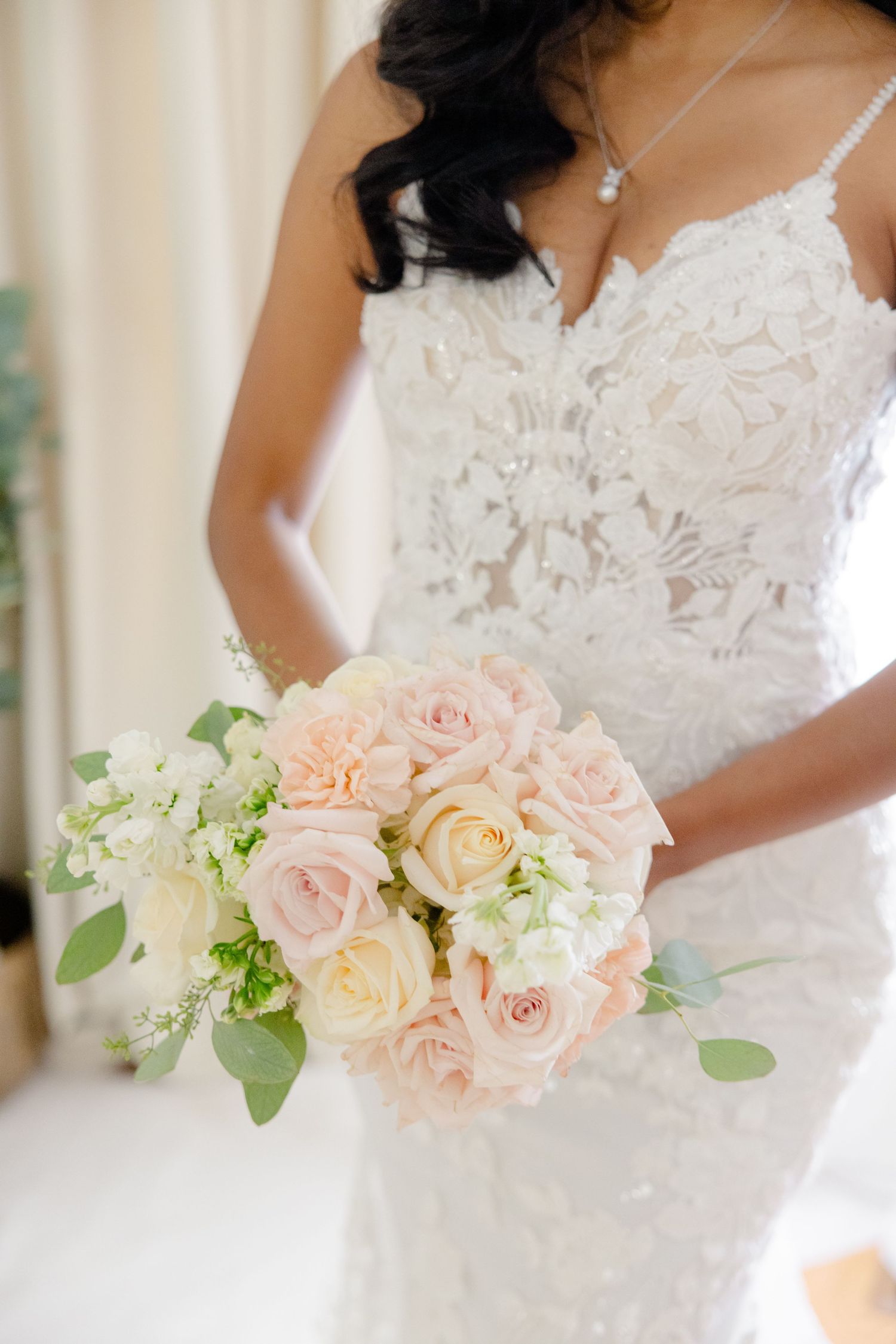 Close up of a romantic pastel bouquet with blush roses and white flowers against a lace wedding dress.