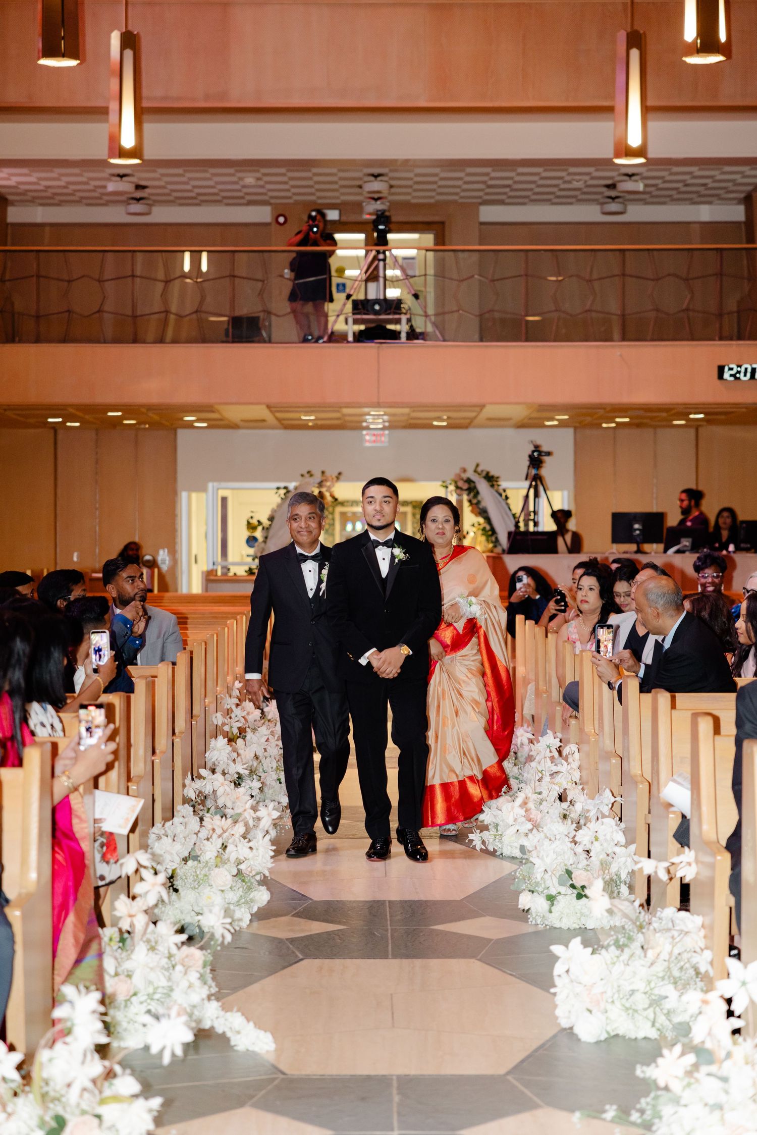 Wedding processional walks down church aisle decorated with white floral arrangements and uplighting.