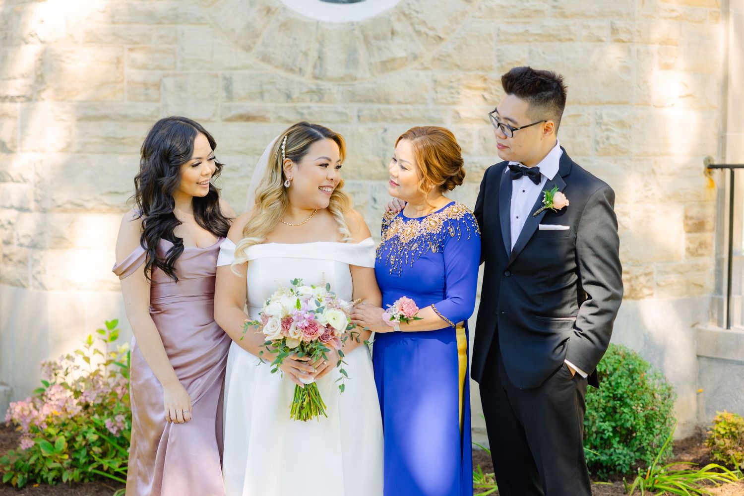Wedding party shares candid moment while posing against stone church wall with circular window.