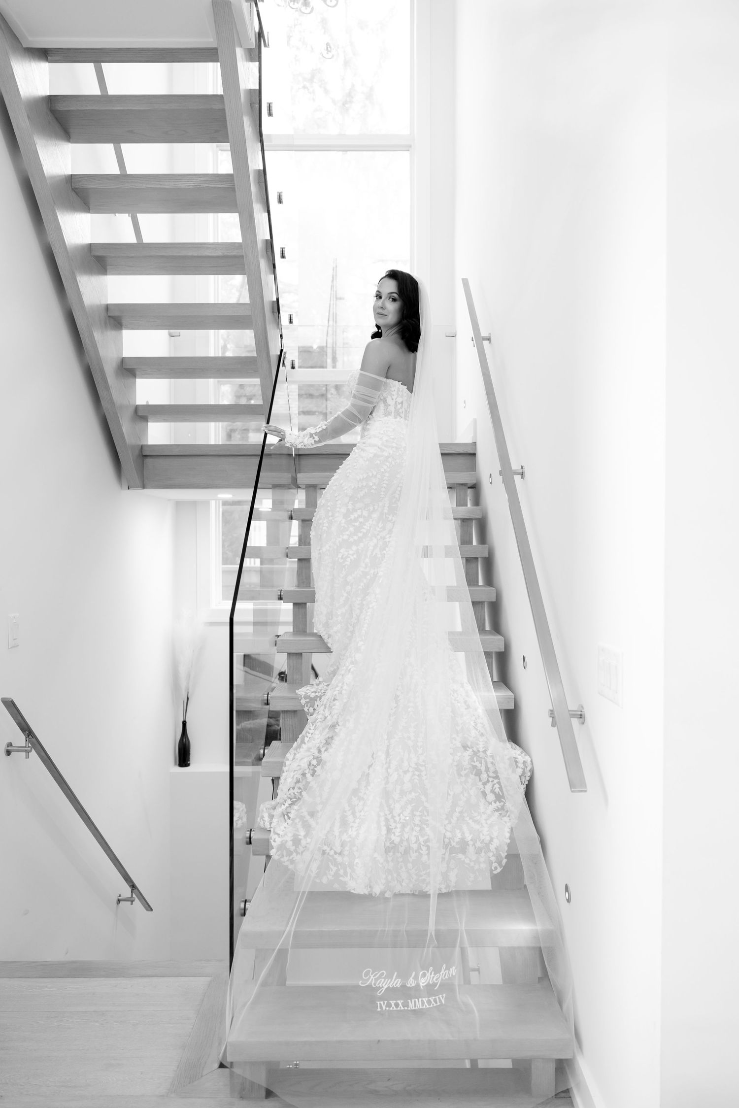Black and white photo of a bride on modern wooden stairs with glass panels.