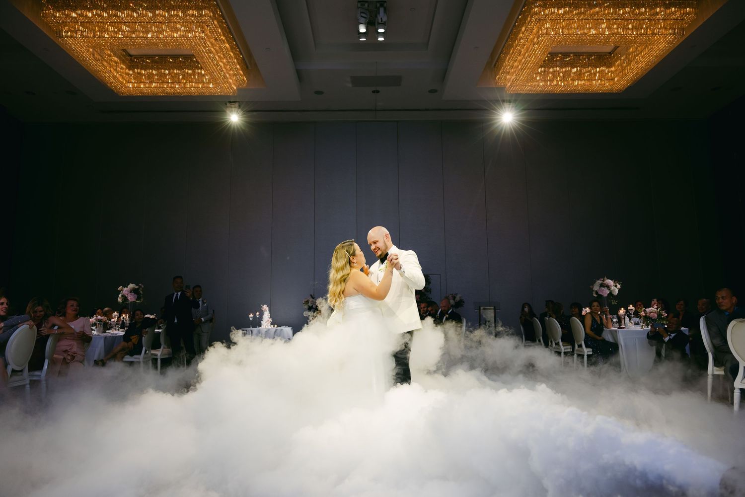 Couple performs first dance amid swirling fog effects under ornate ceiling lights.