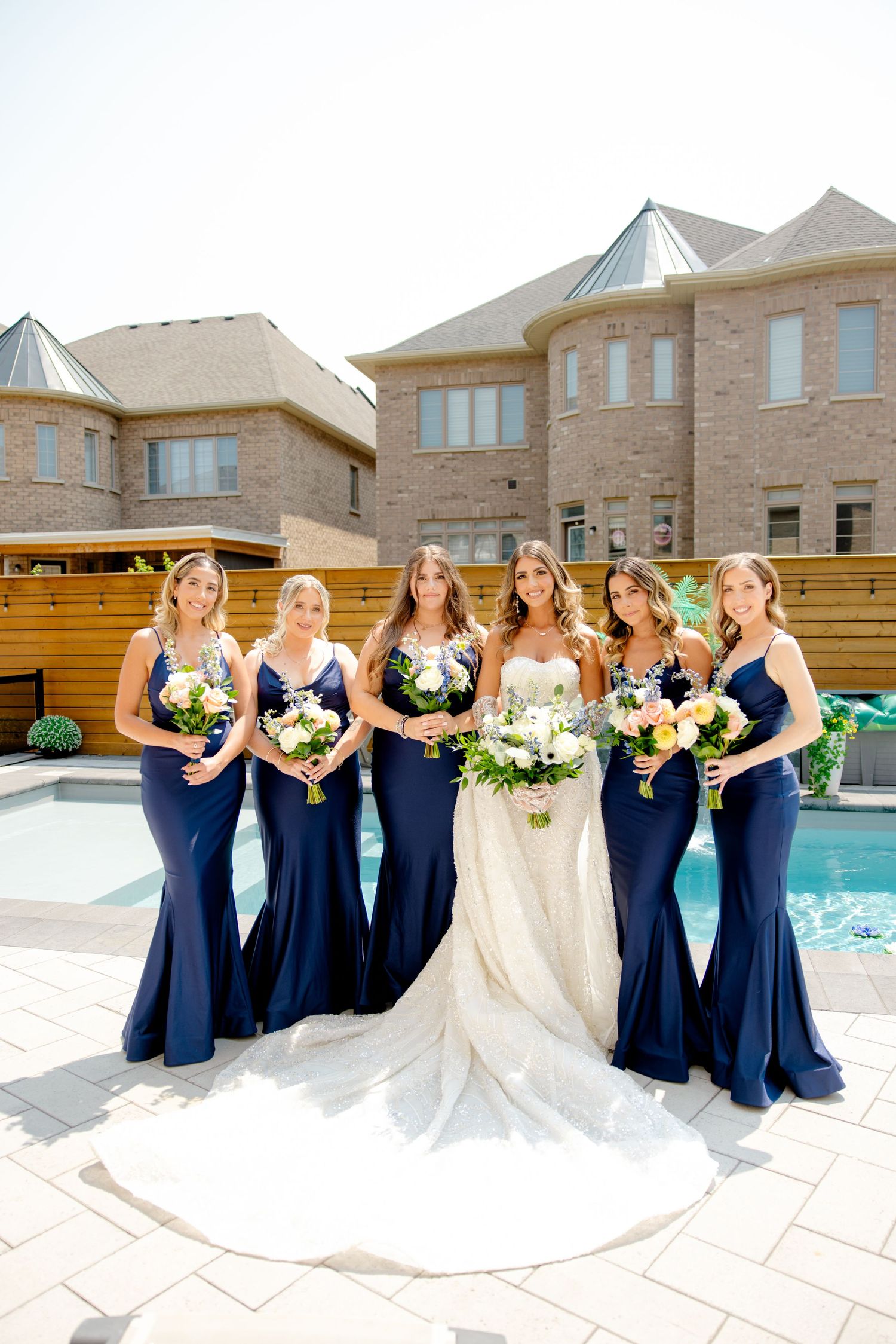 Wedding party in navy blue bridesmaid dresses posing by a pool with elegant bouquets.