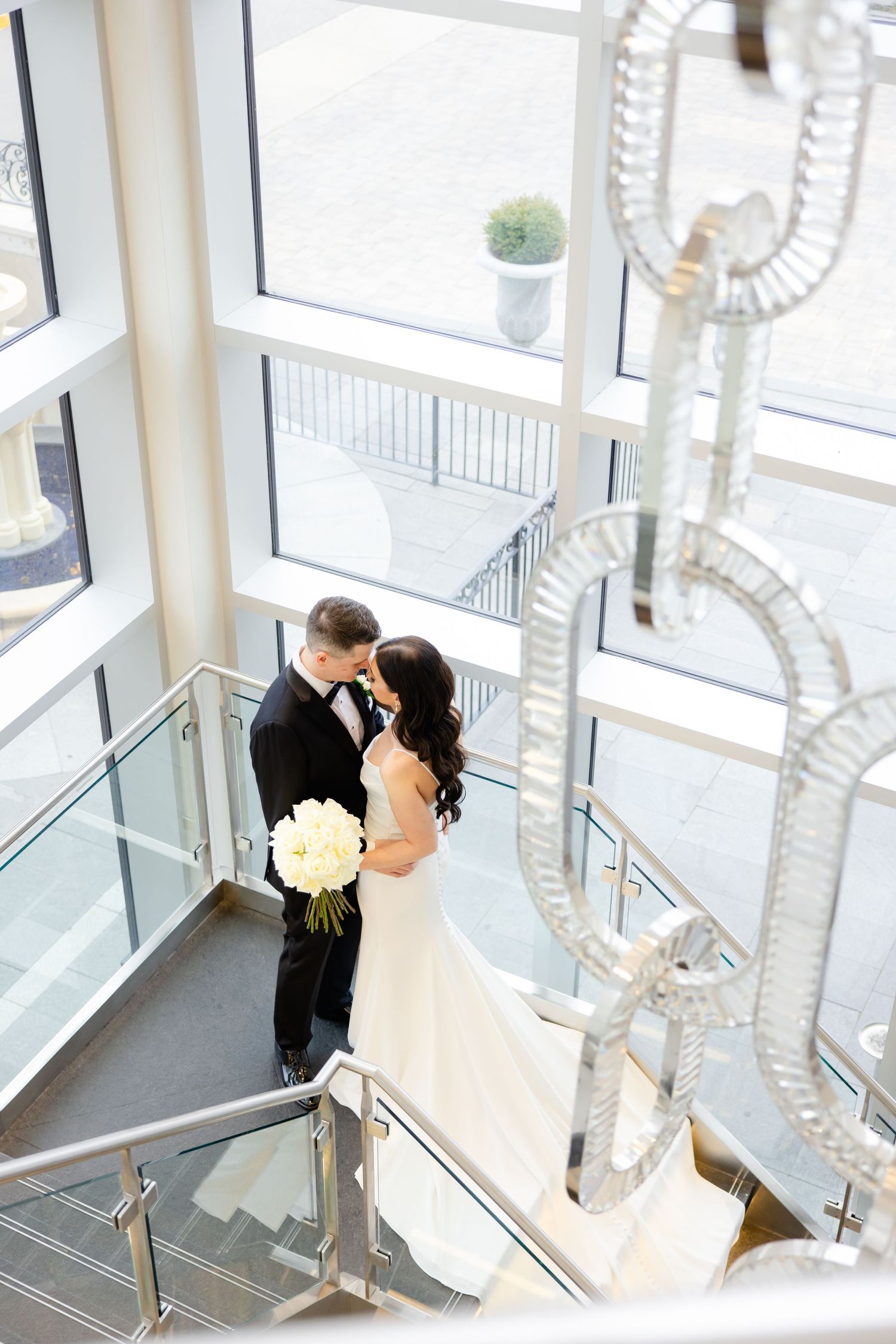 Romantic wedding moment captured in modern stairwell with large windows and chandelier.