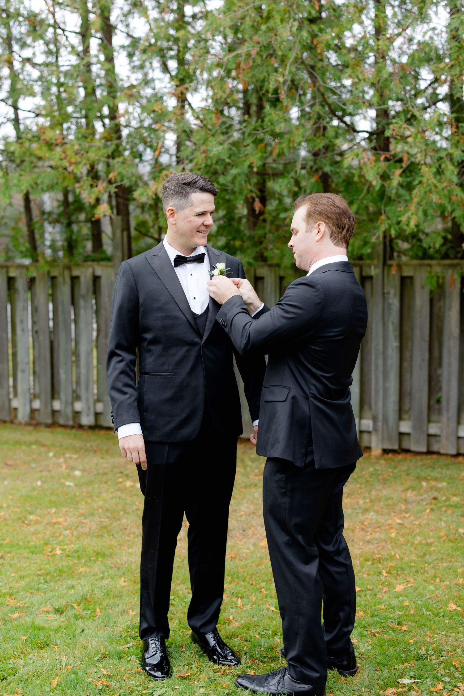 Two people in formal attire adjust a boutonniere on a suit lapel in a garden setting.
