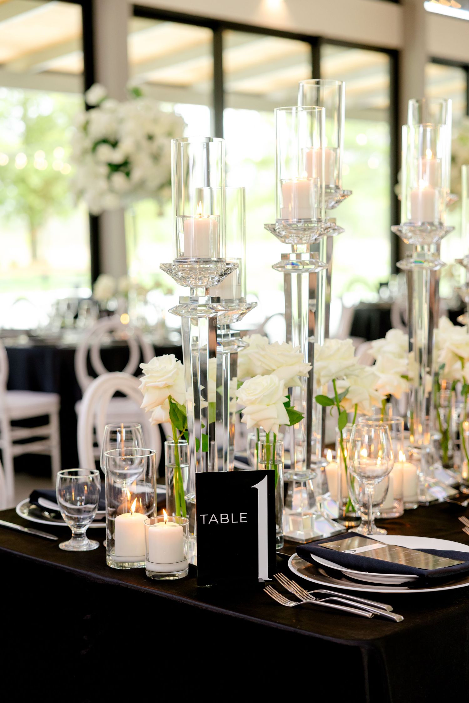 Modern wedding table setting with tall glass candleholders, white florals, and polished silverware.