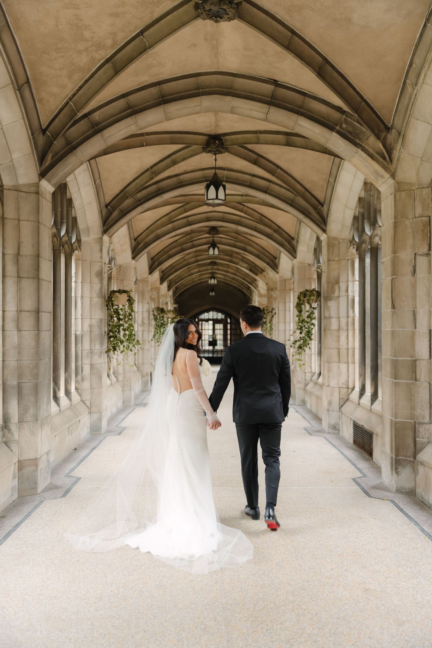 Wedding couple walks through an arched stone hallway decorated with floral arrangements and pendant lights.