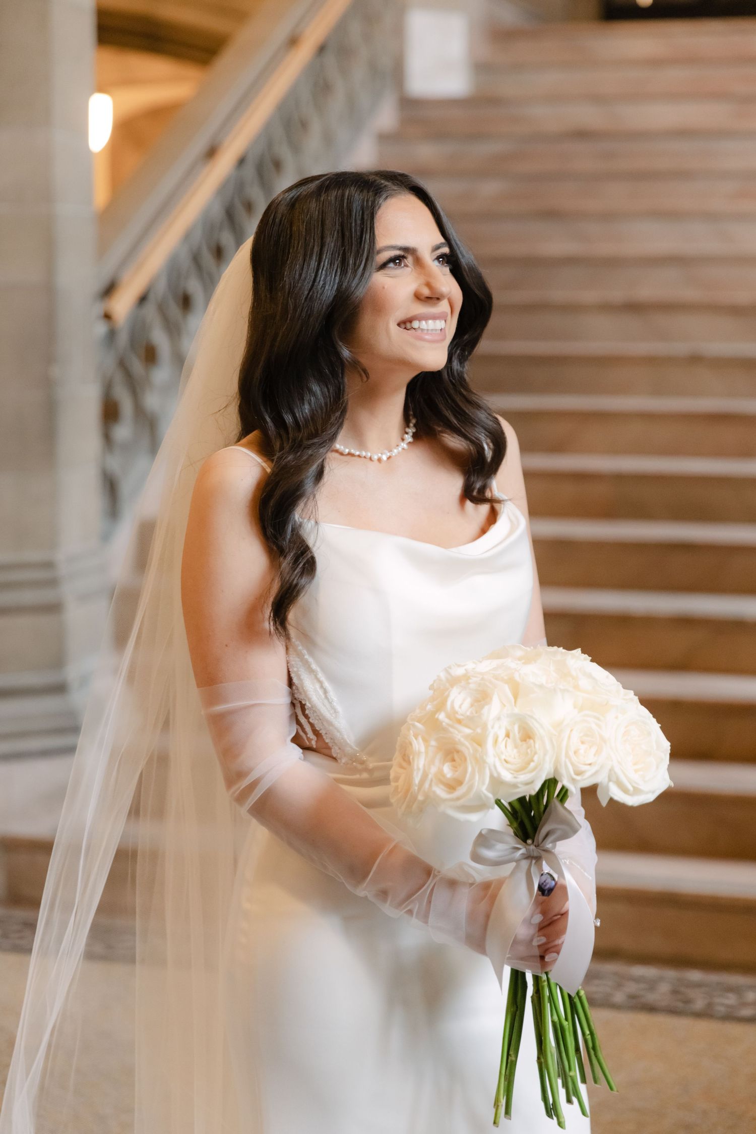 A bridal portrait showcasing an elegant white gown and bouquet of cream roses on classic marble stairs.
