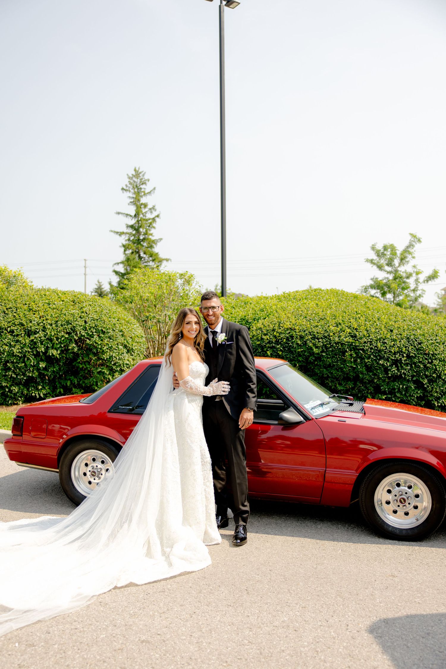 Wedding couple poses beside classic red sports car with manicured hedges in background.
