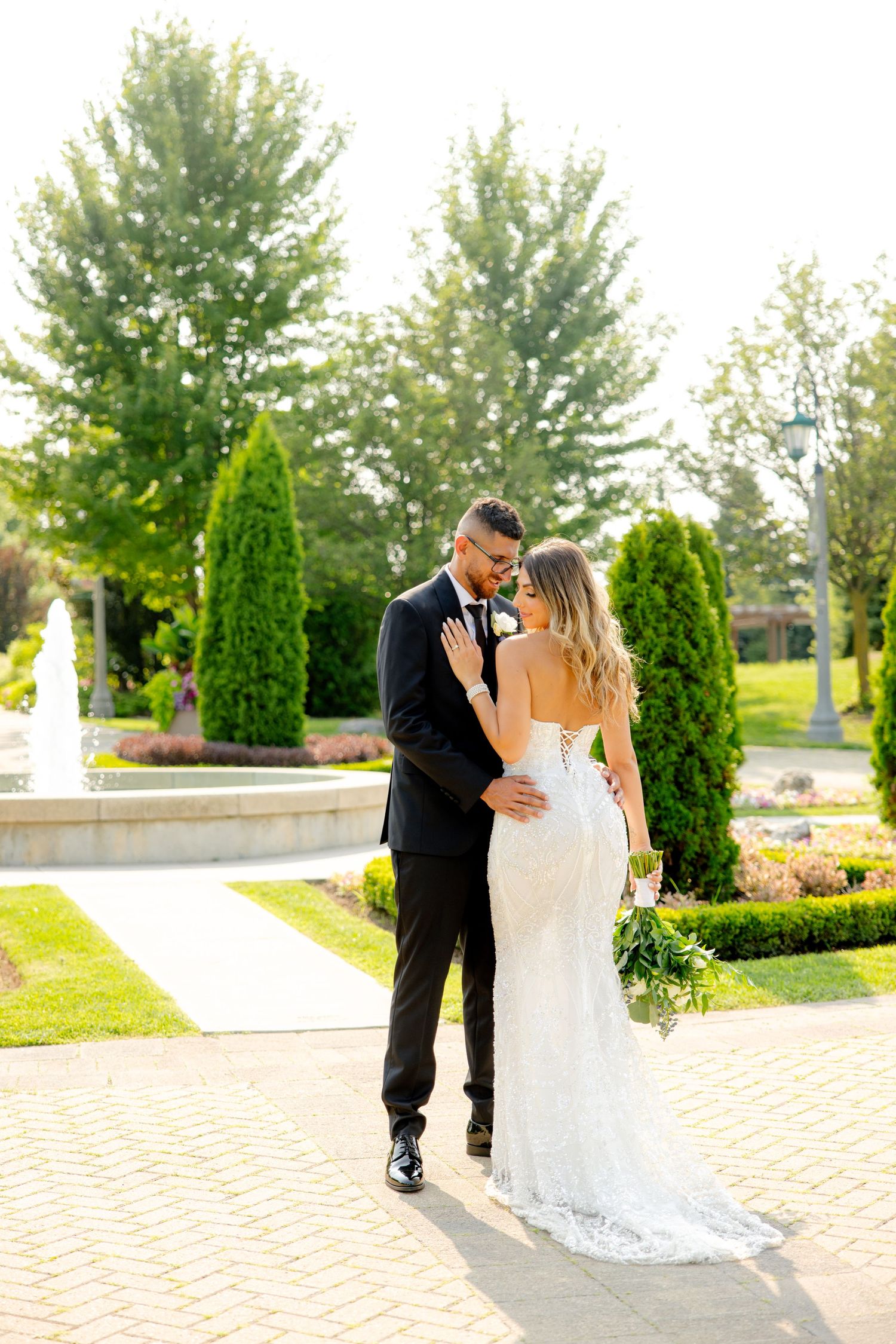 Wedding couple sharing tender moment on ornate garden grounds with fountain and topiary trees.