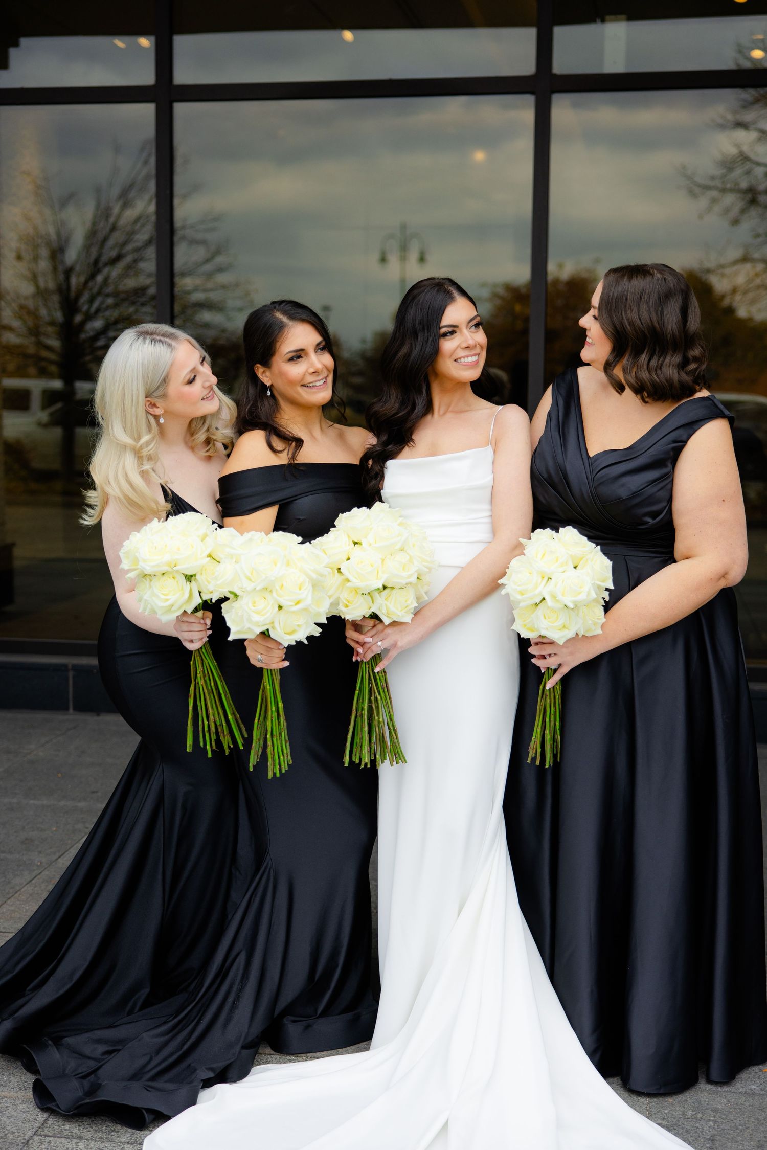 Bridesmaids in black gowns gather around bride in white, all holding cream-colored bouquets outdoors.