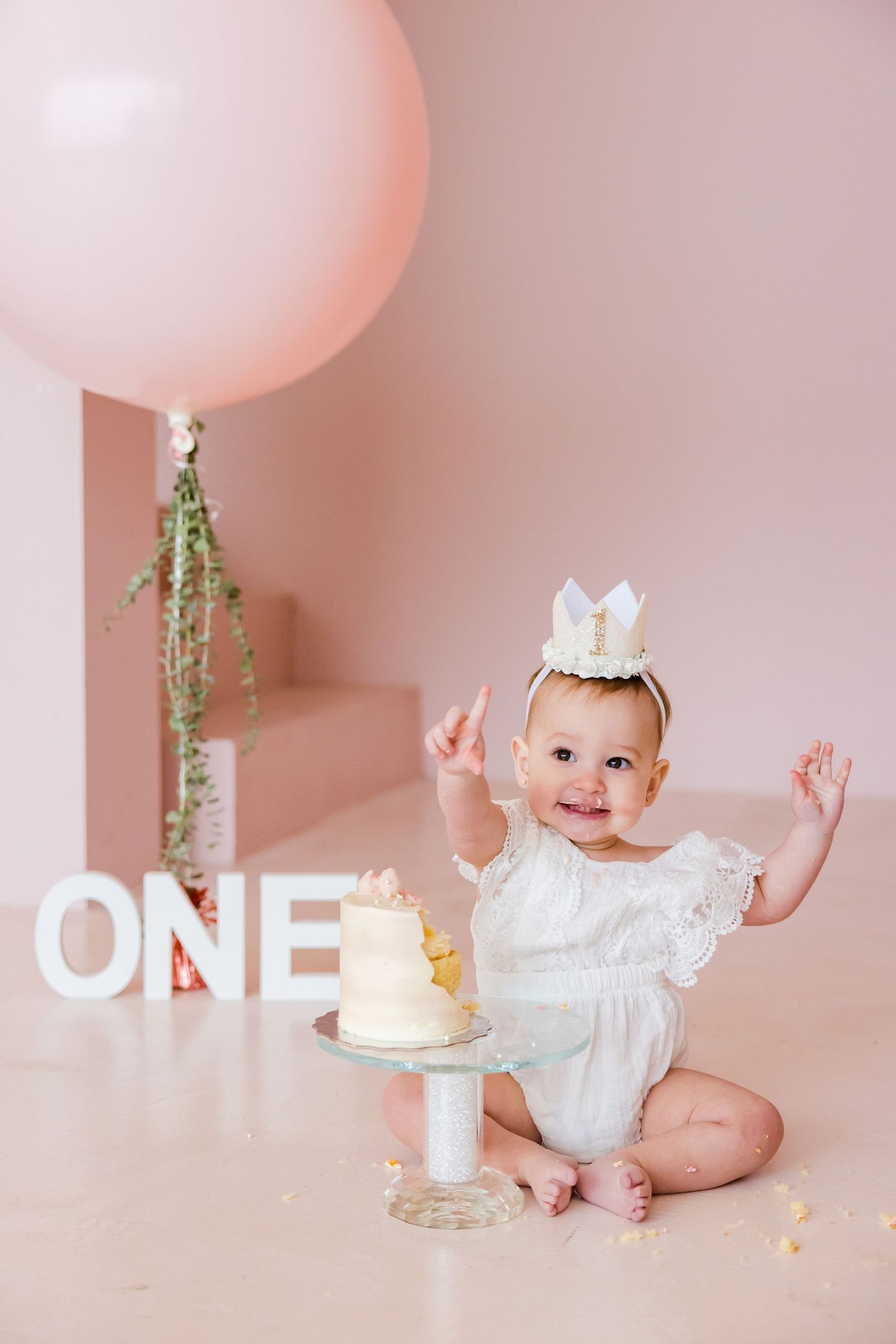 First birthday portrait with white cake, pink balloon, and delicate crown.