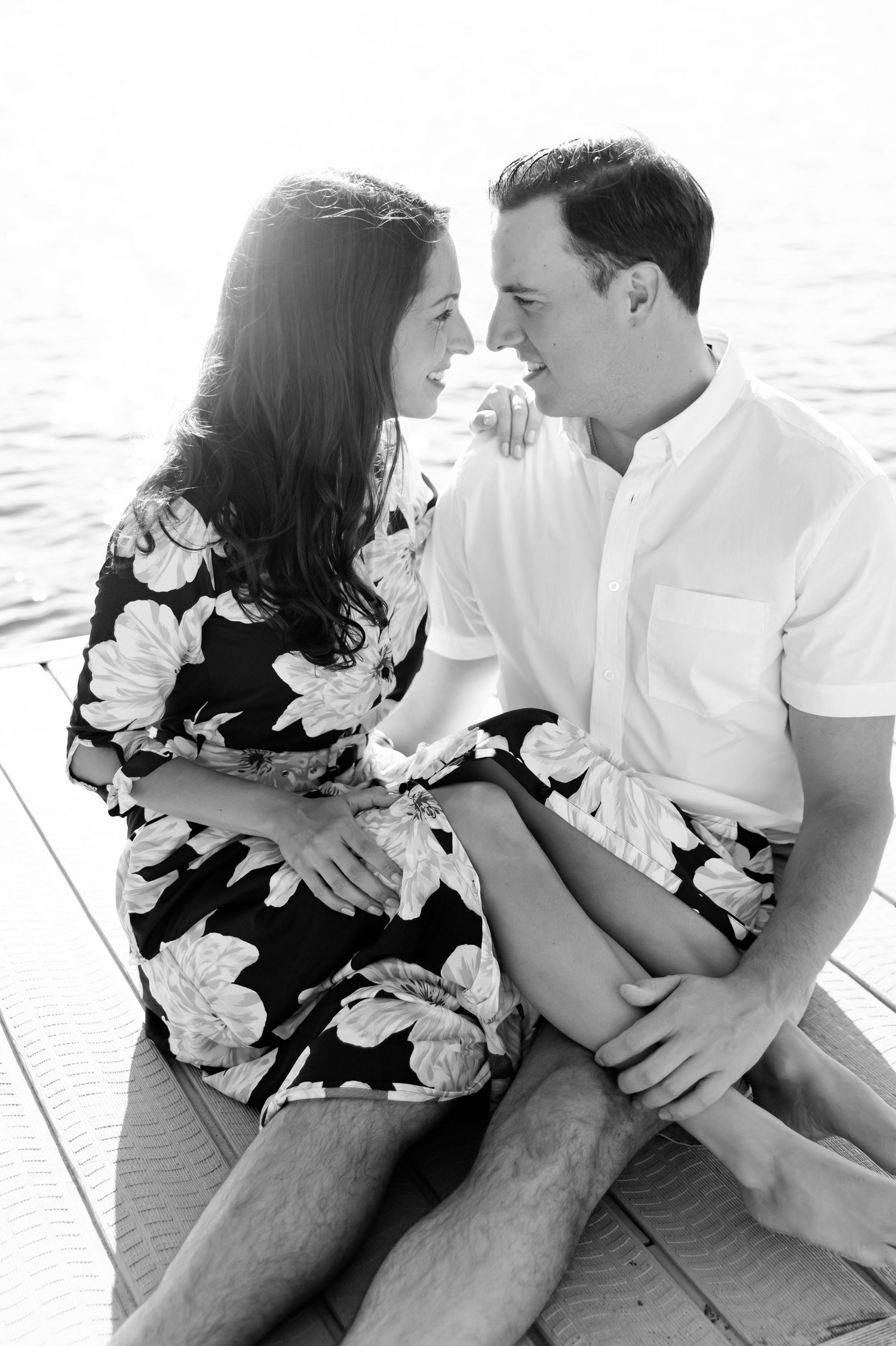 An intimate black and white portrait of a couple sitting close together on a lakeside dock.