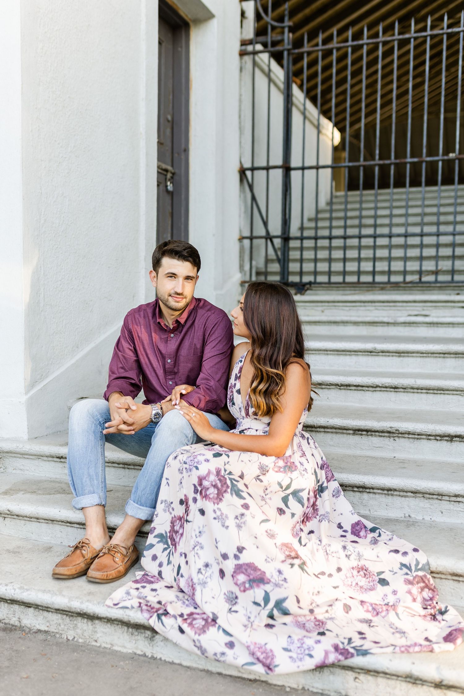 Couple sitting on concrete steps, wearing purple shirt and floral dress during outdoor photoshoot.