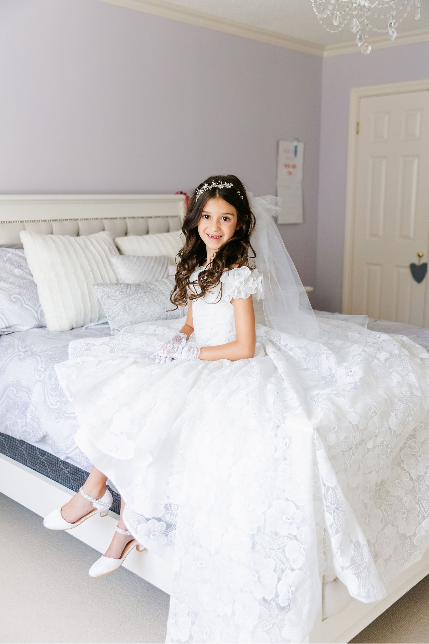 White bedroom setting with elegant wedding dress displayed on plush bedding under crystal chandelier.