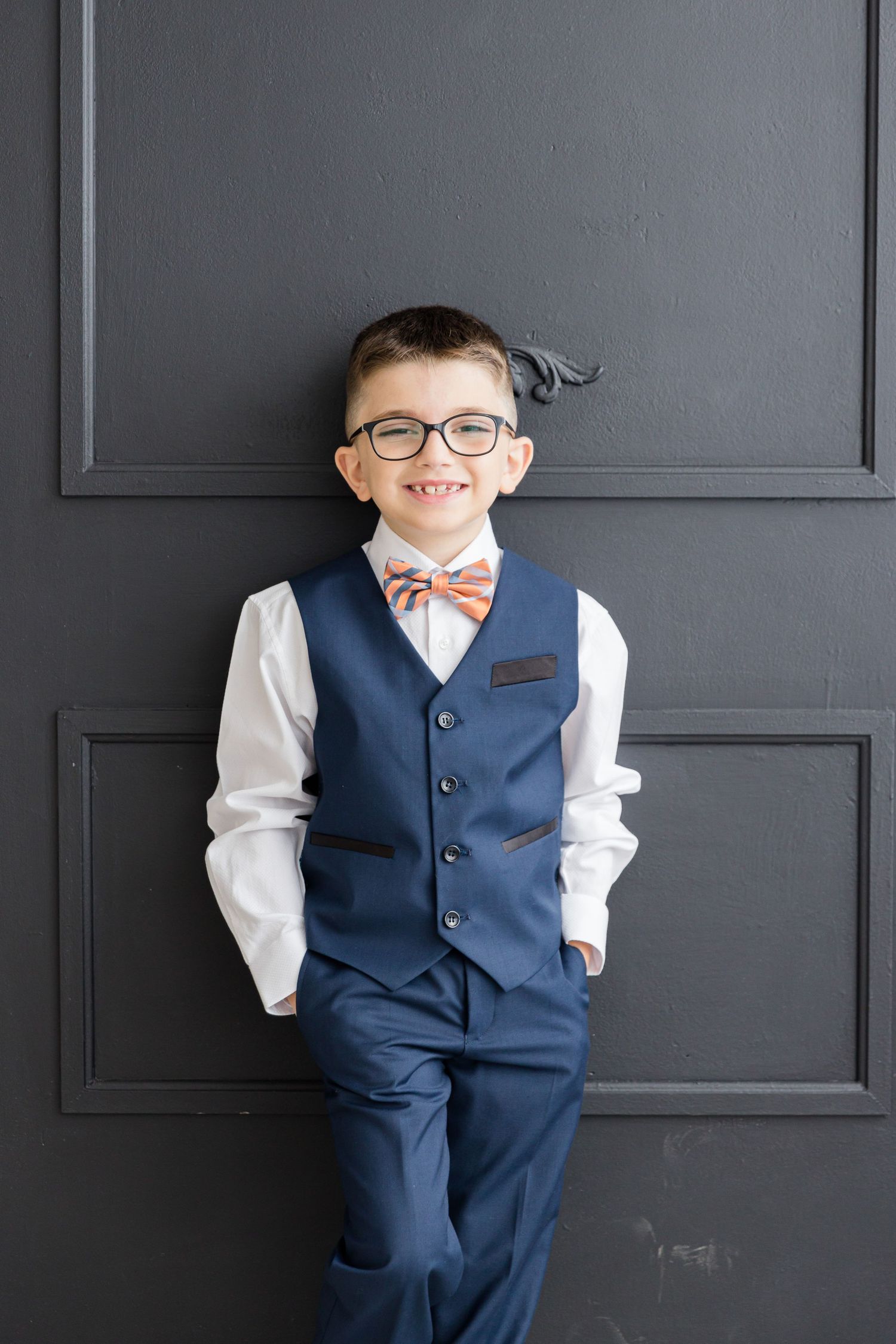 Smart formal attire with glasses and peach bow tie against a dark paneled wall.