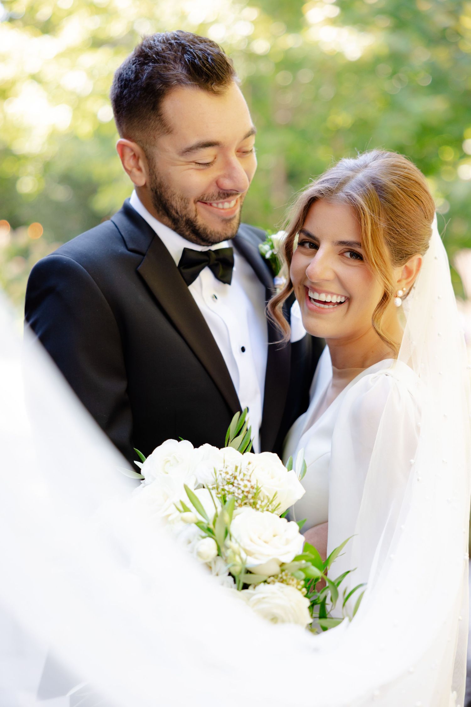 Joyful wedding portrait with flowing veil and white bouquet in natural outdoor setting.