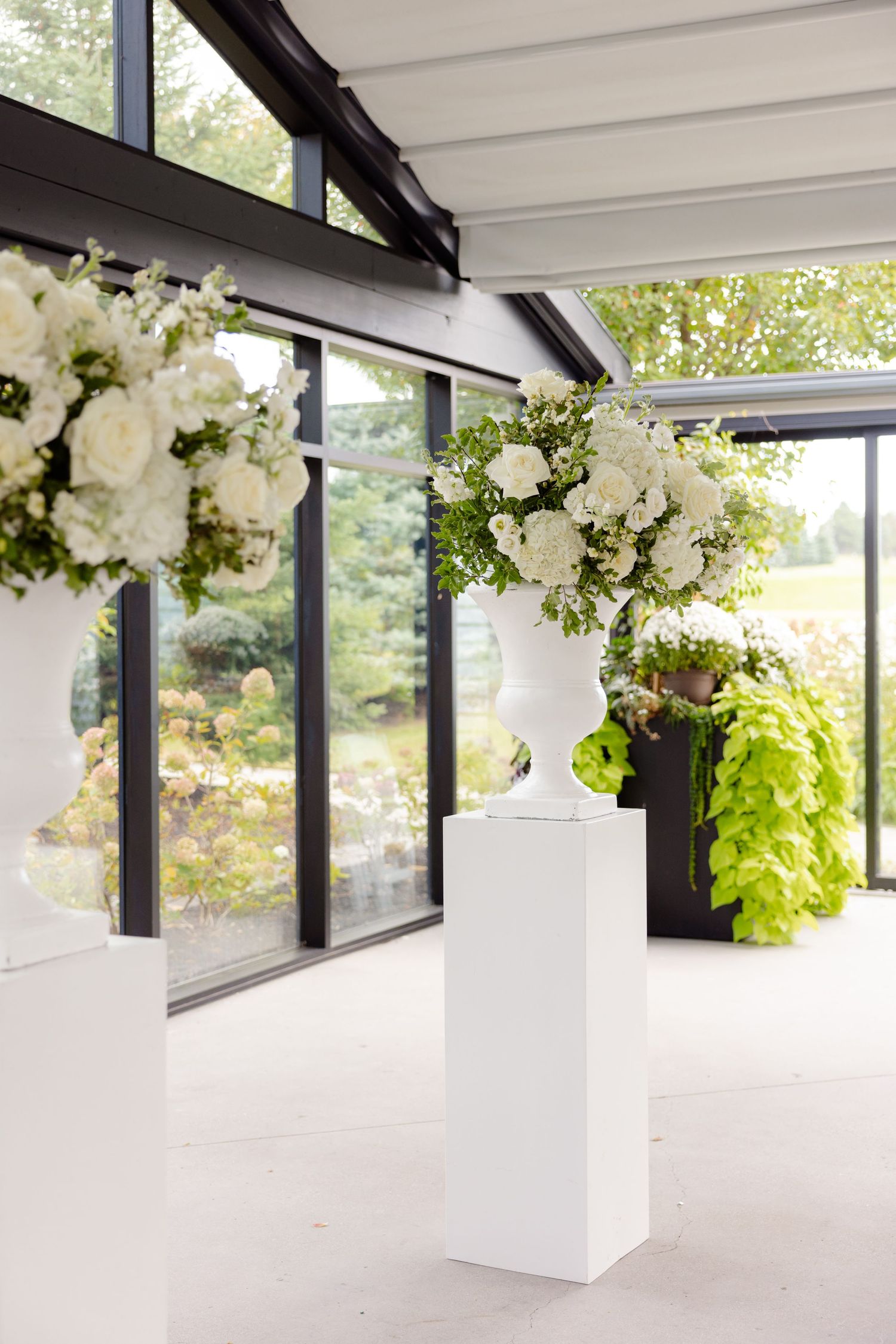 Elegant white floral arrangements on pedestals decorate a bright indoor venue with floor-to-ceiling windows.
