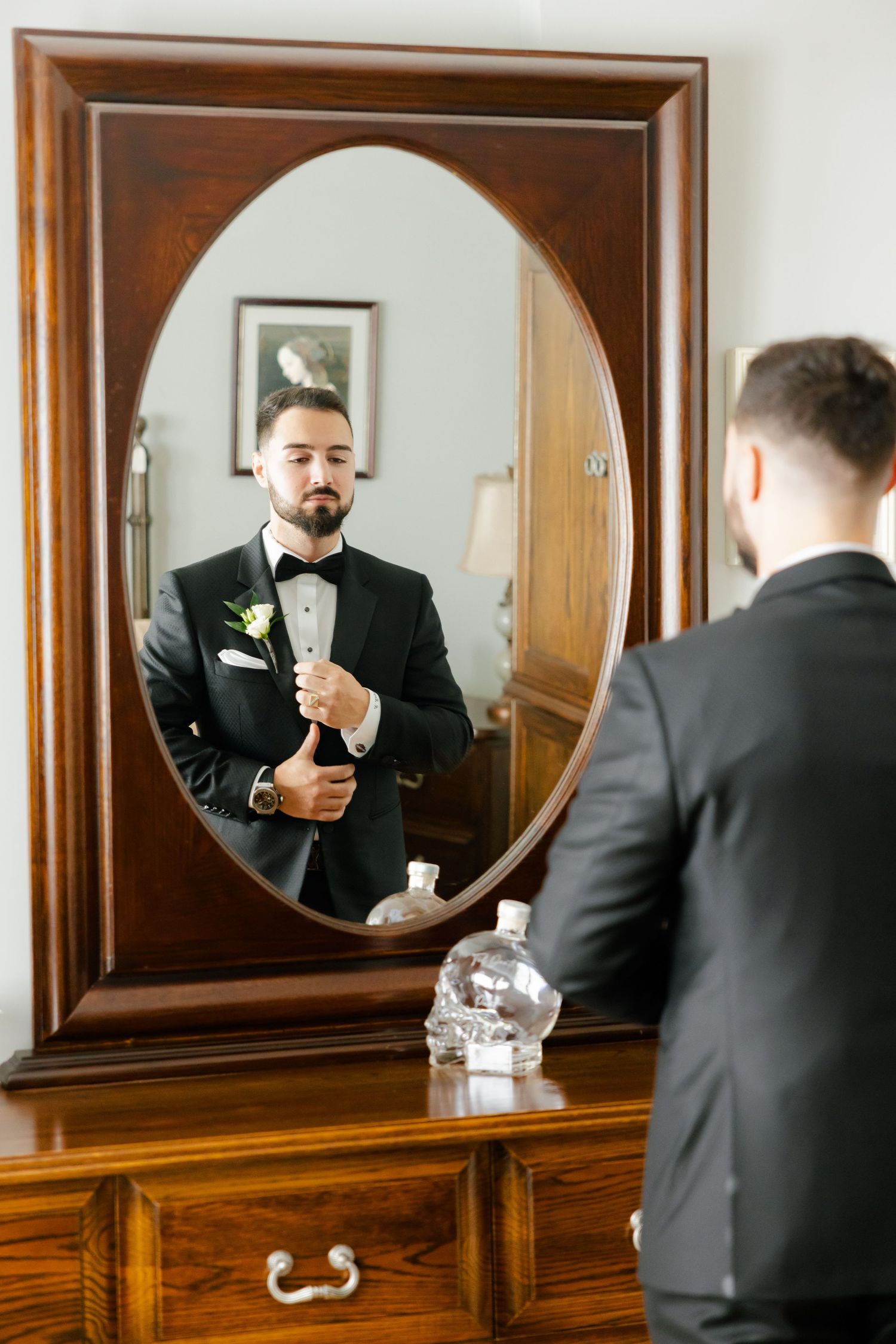 A groom in a black tuxedo adjusts his cufflinks while looking in an oval mirror atop a wooden dresser.