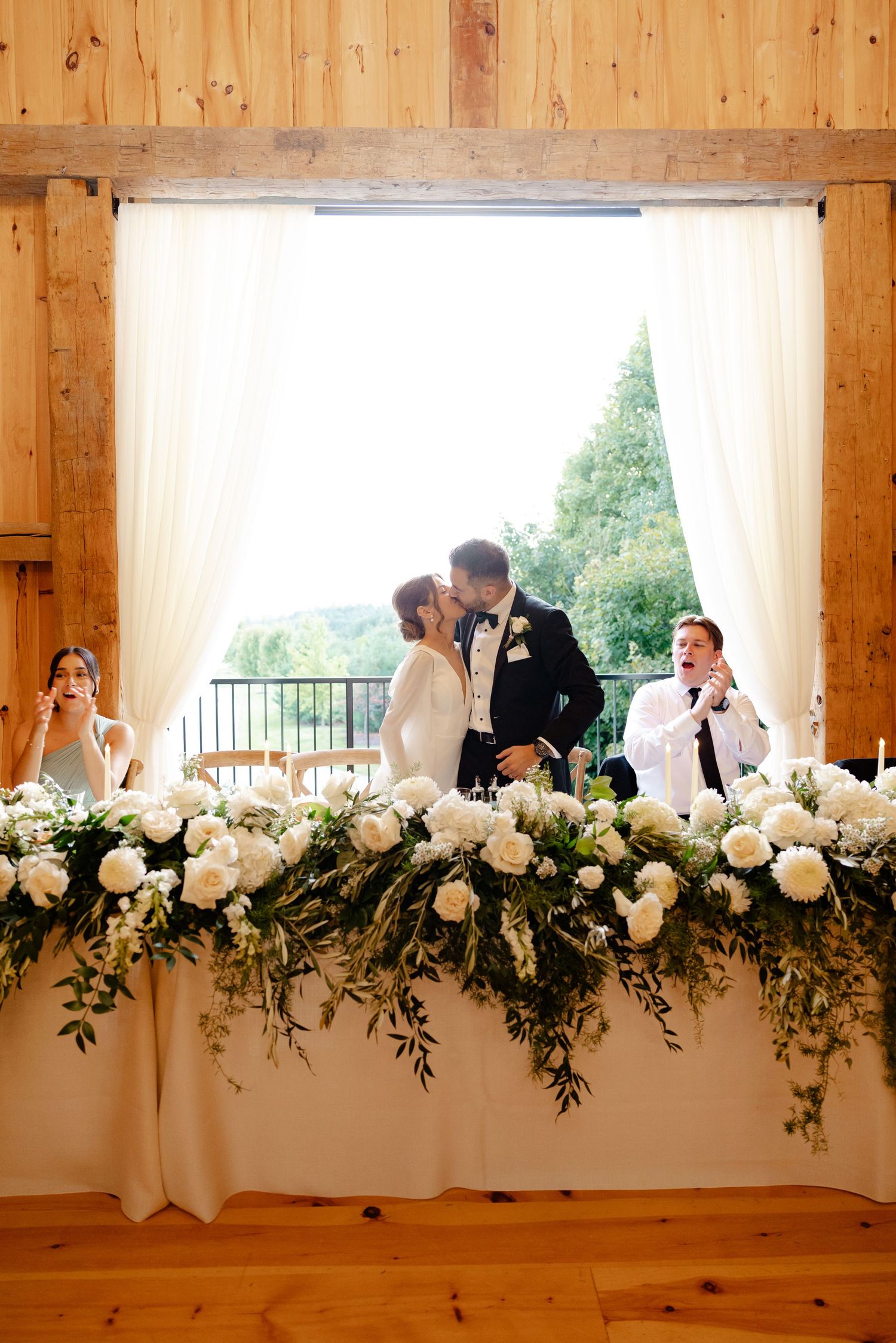 A romantic moment shared at a wedding head table decorated with lush white floral arrangements in a rustic barn venue.