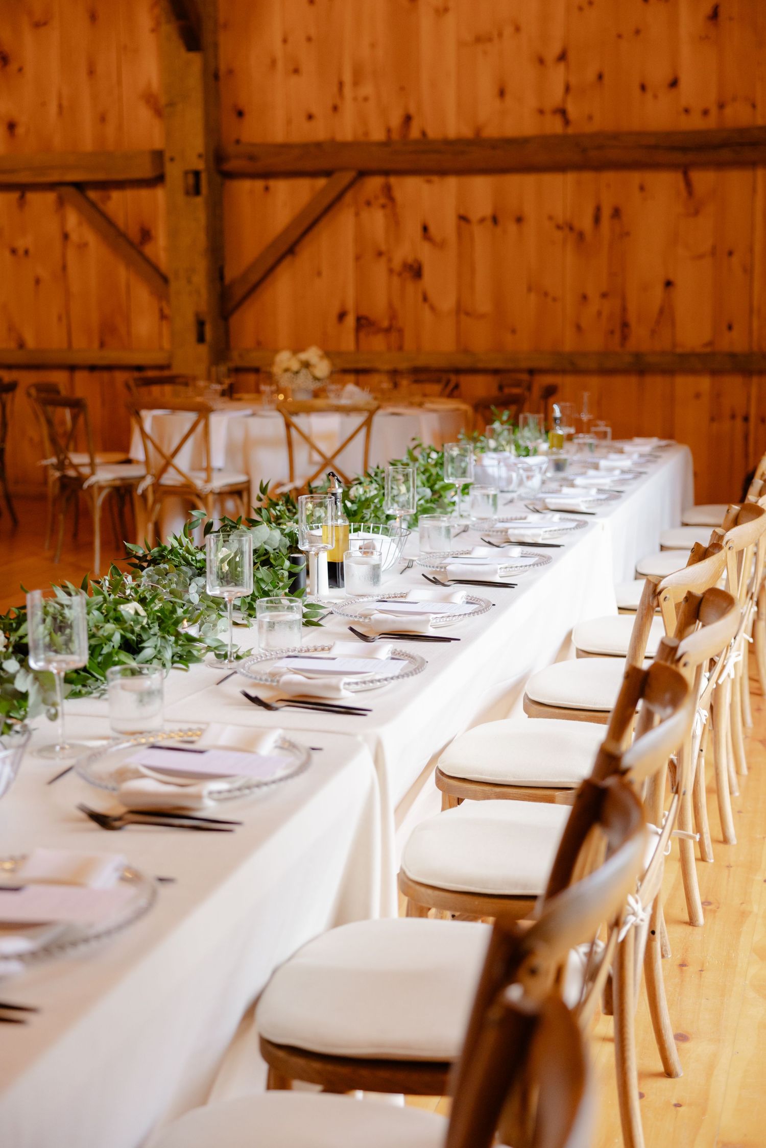 Long banquet table with white linens and crossback chairs decorated with flowing greenery centerpieces.