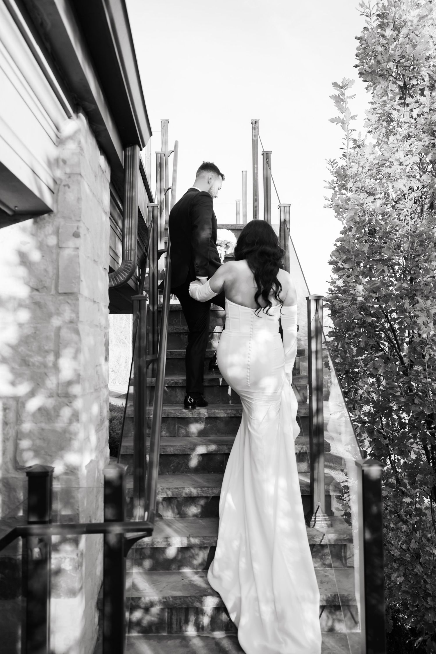 Black and white photo of newlyweds ascending outdoor stairs in elegant wedding attire.