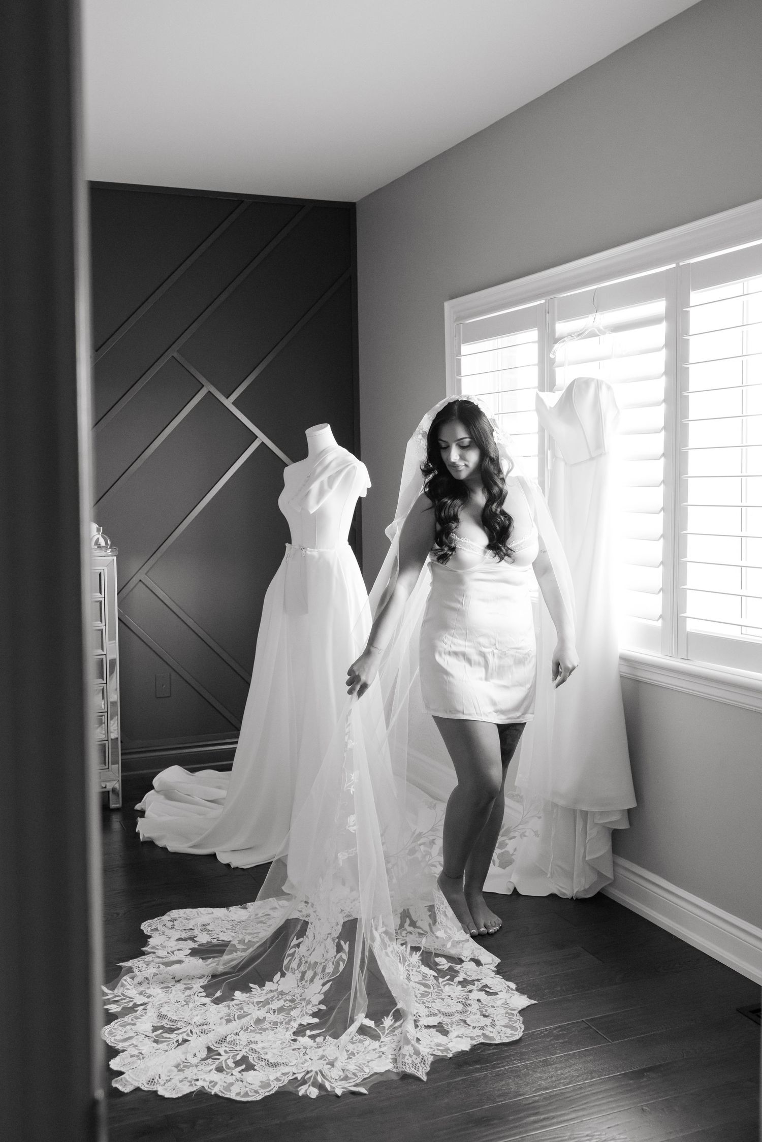 Black and white photo of bride in short dress standing next to wedding gown on display.